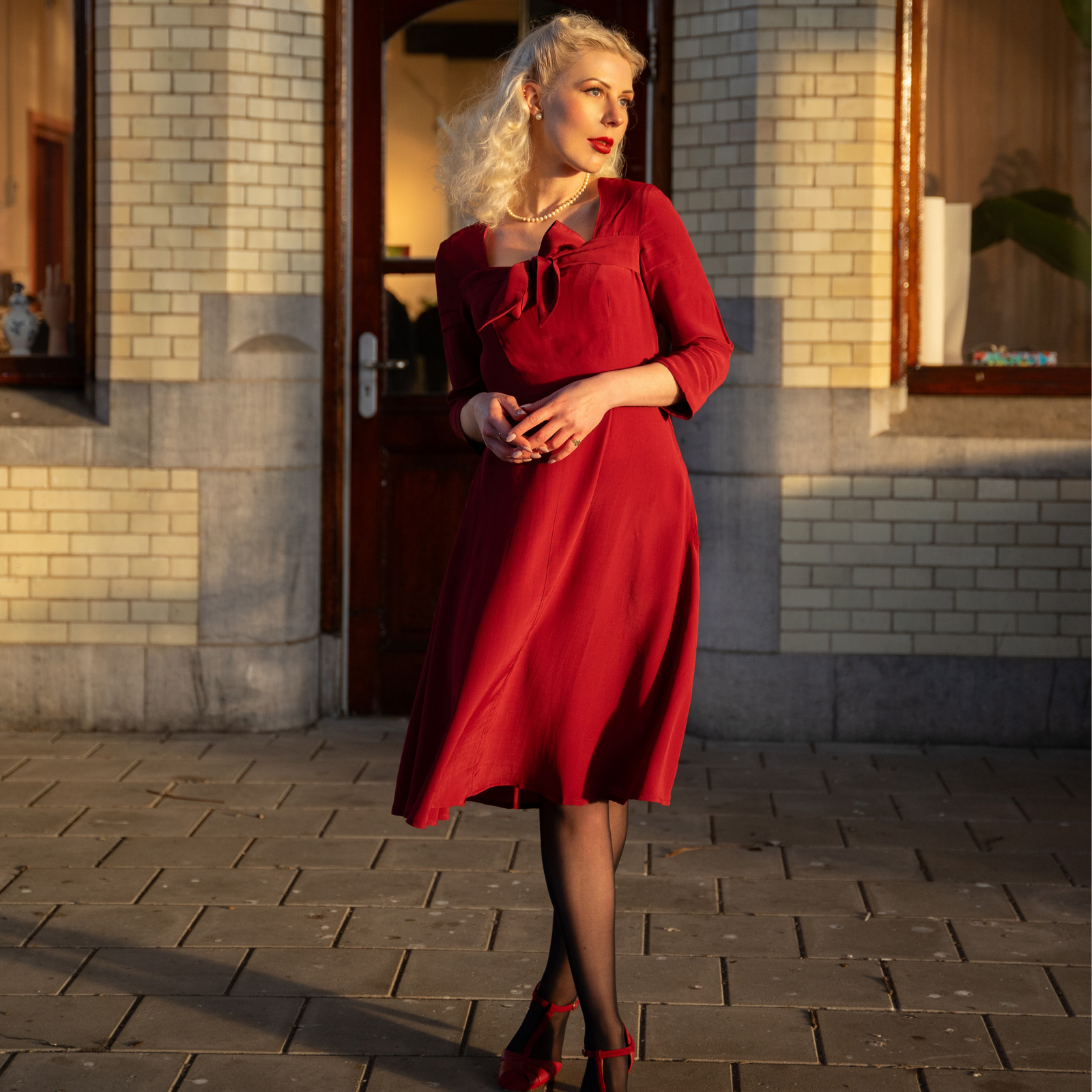 A woman standing outside wearing a red, long-sleeved, knee-length dress with a sweetheart neckline and a center front pussy bow.