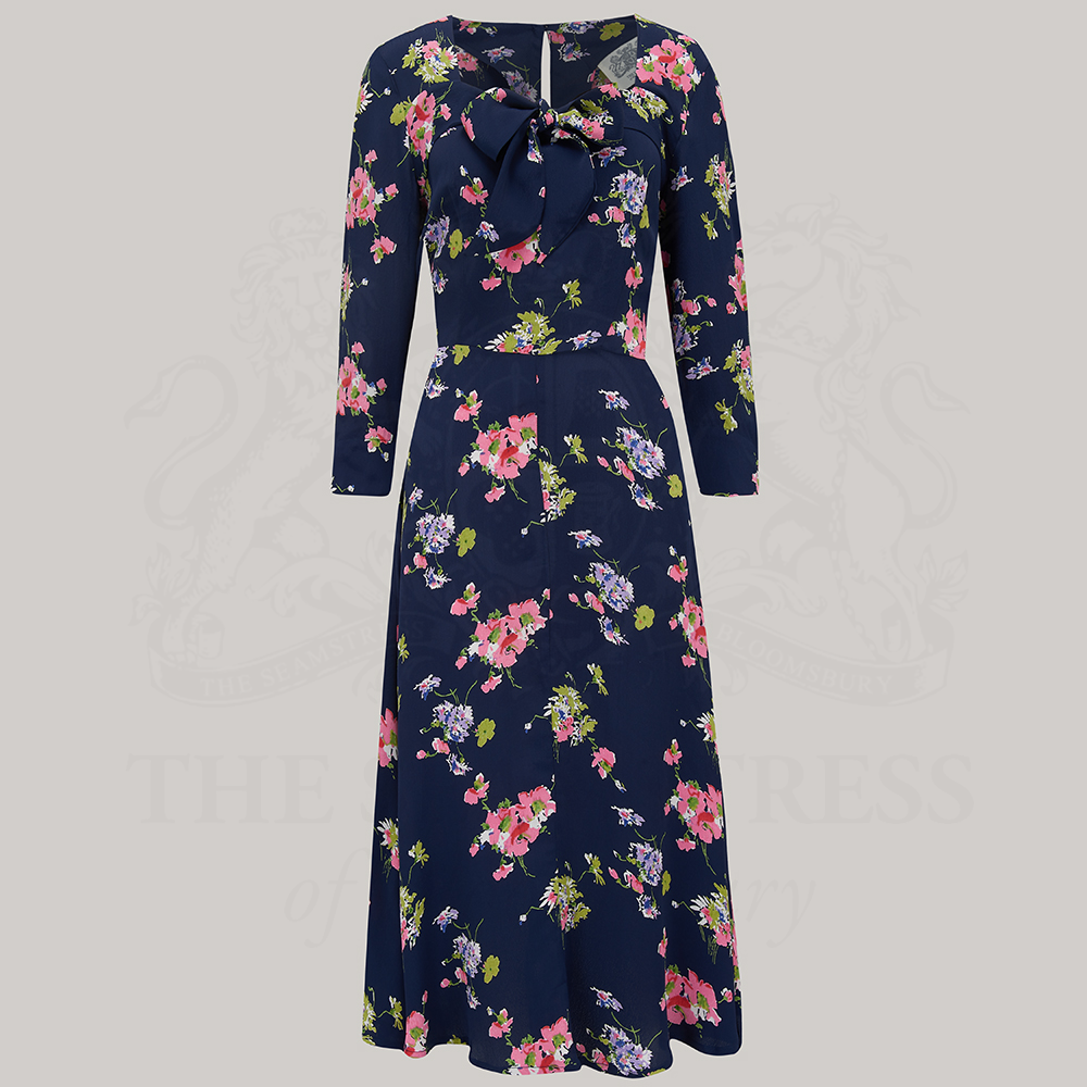 This image features the Joyce Dress in Navy Mayflower, exuding elegance and style from its inception in the 1940s. The dress is presented in a rich navy blue color, adorned with a vibrant floral pattern