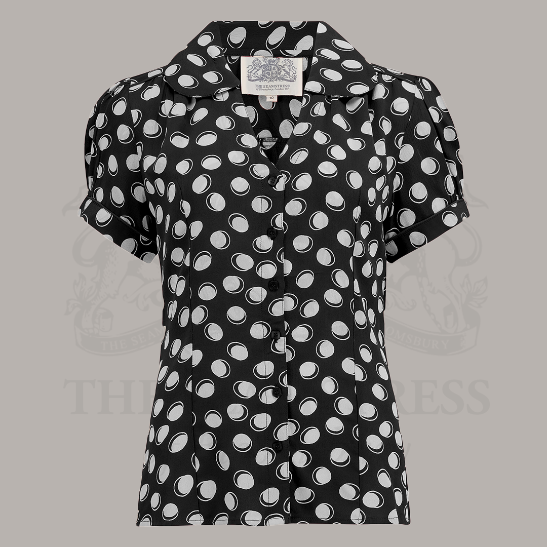 A vintage-style black blouse with white polka dots, featuring a Peter Pan-style collar and puff sleeves.