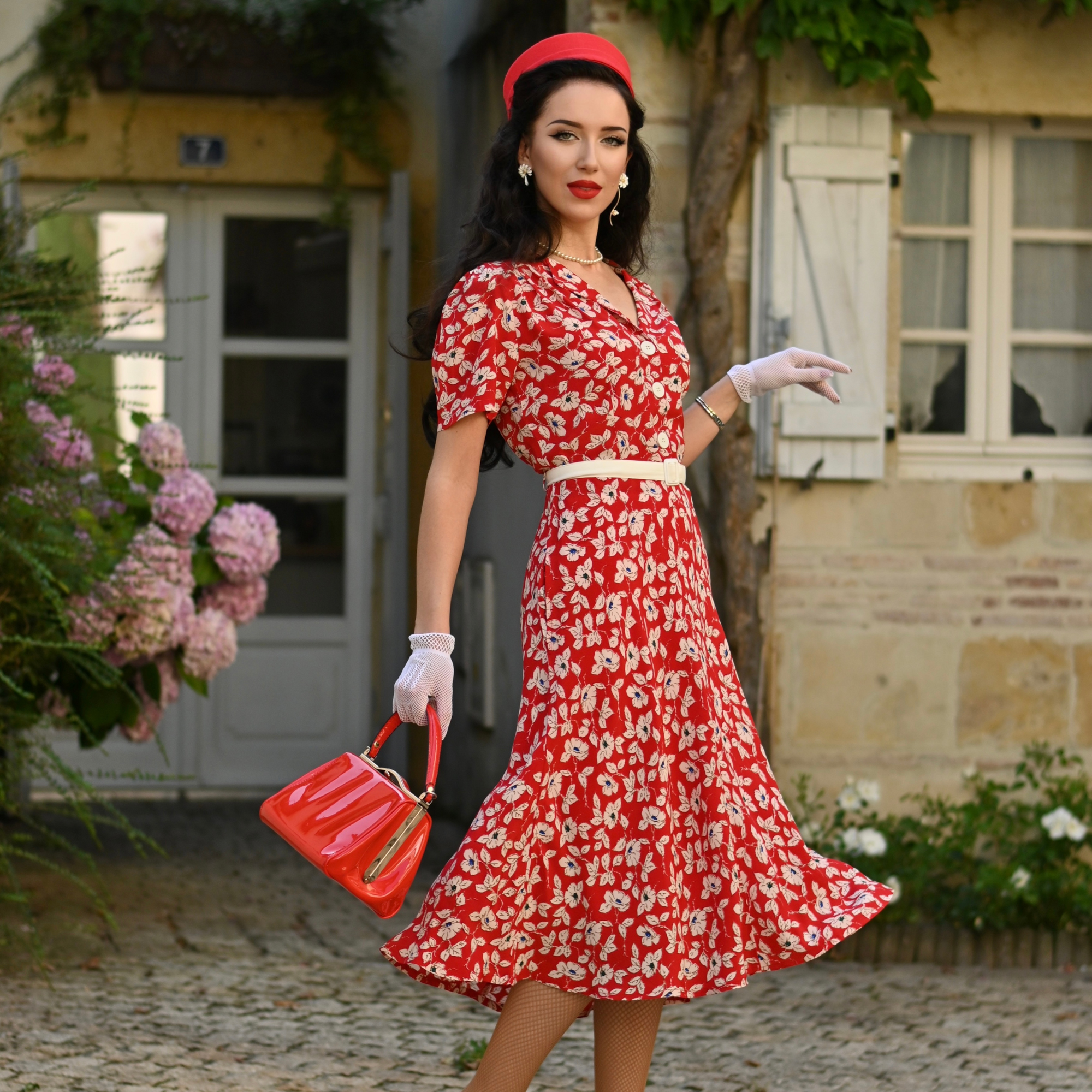 A woman standing in front of a house wearing a red shirtwaist dress with a pansy print, white gloves, and a red hat. She is carrying a red handbag.