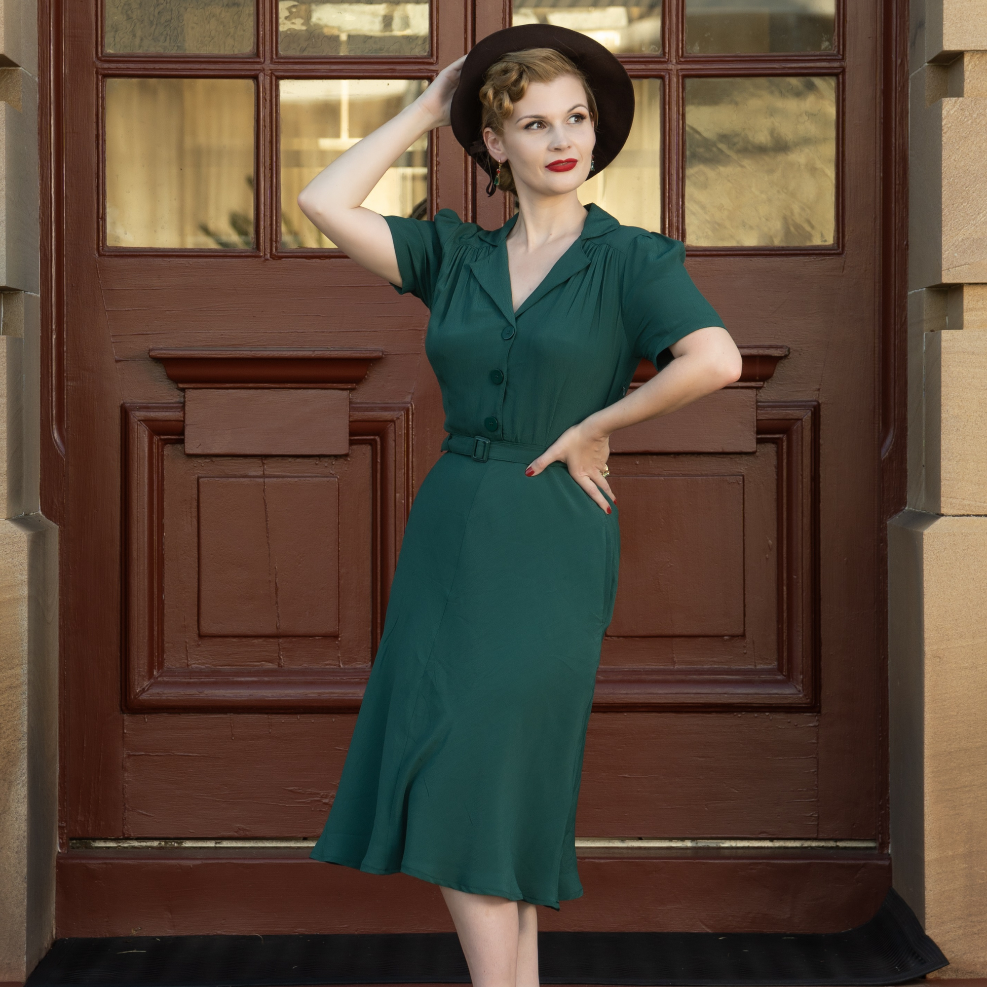 A woman standing in front of a dark door wearing a green, tailored, shirtwaist style dress with a button-down bodice and a matching waist belt. She has a hat on and the dress has side-seam pockets and a hidden side zip.