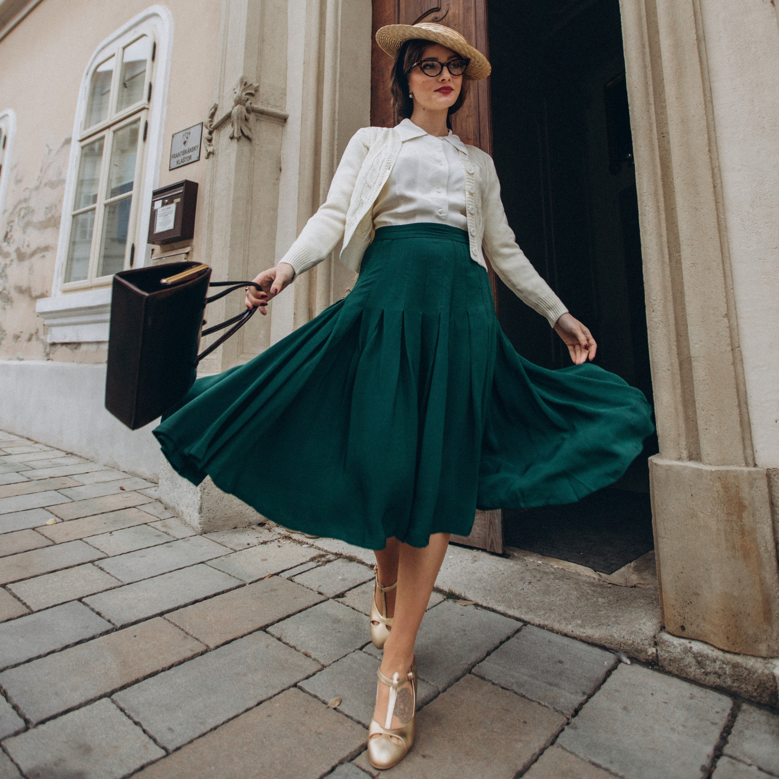 A person wearing a vintage style pleated skirt in green, with a cream blouse and a wide-brimmed hat, standing on a cobblestone street.