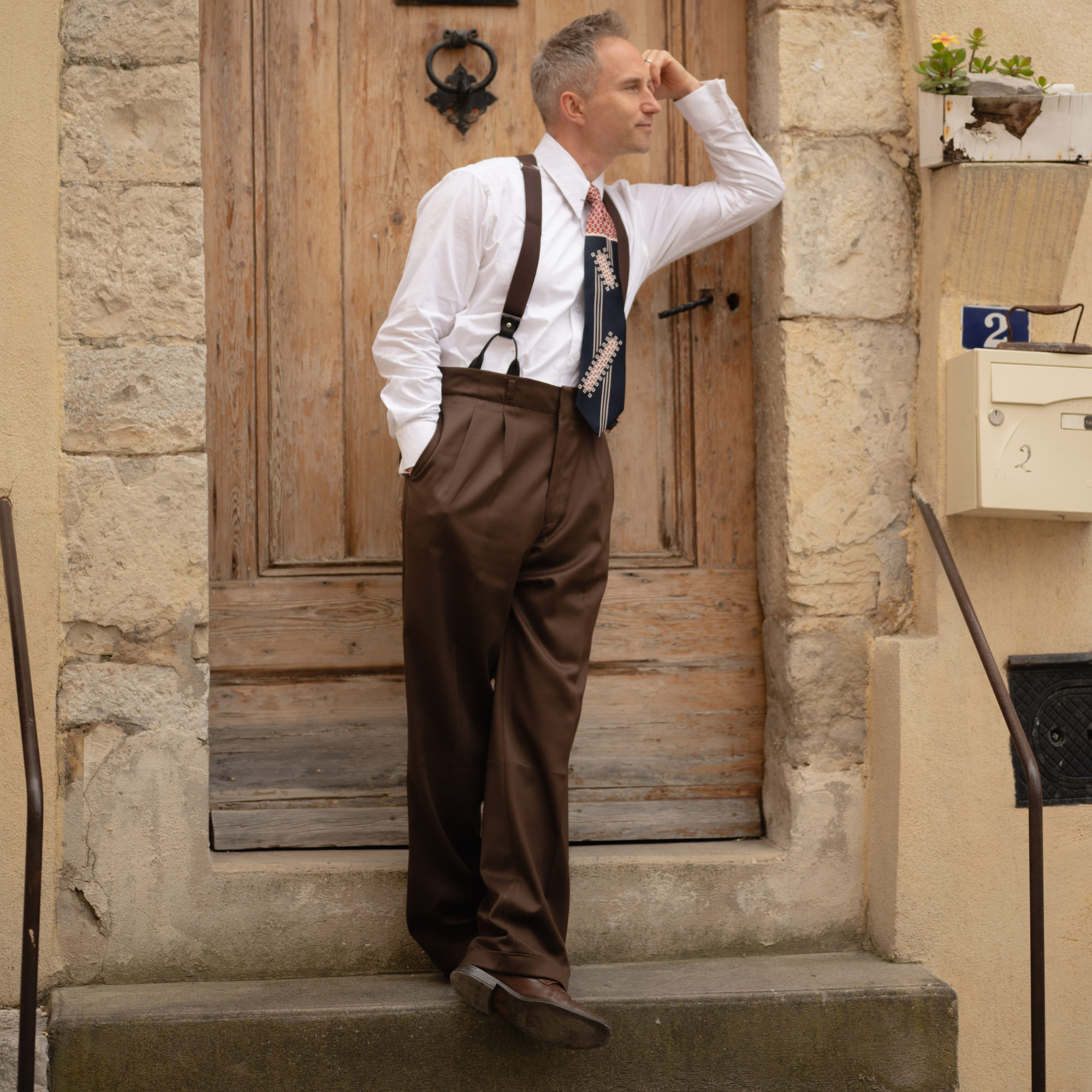 A man wearing a brown pair of men's trousers, paired with a white spearpoint shirt, braces and tie. He stands outside on stone steps.