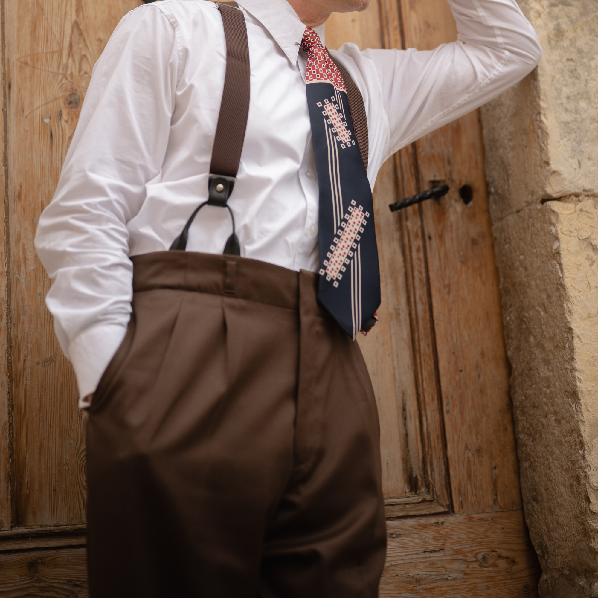 A man wearing a brown pair of men's trousers, paired with a white spearpoint shirt, braces and tie. He stands outside on stone steps.