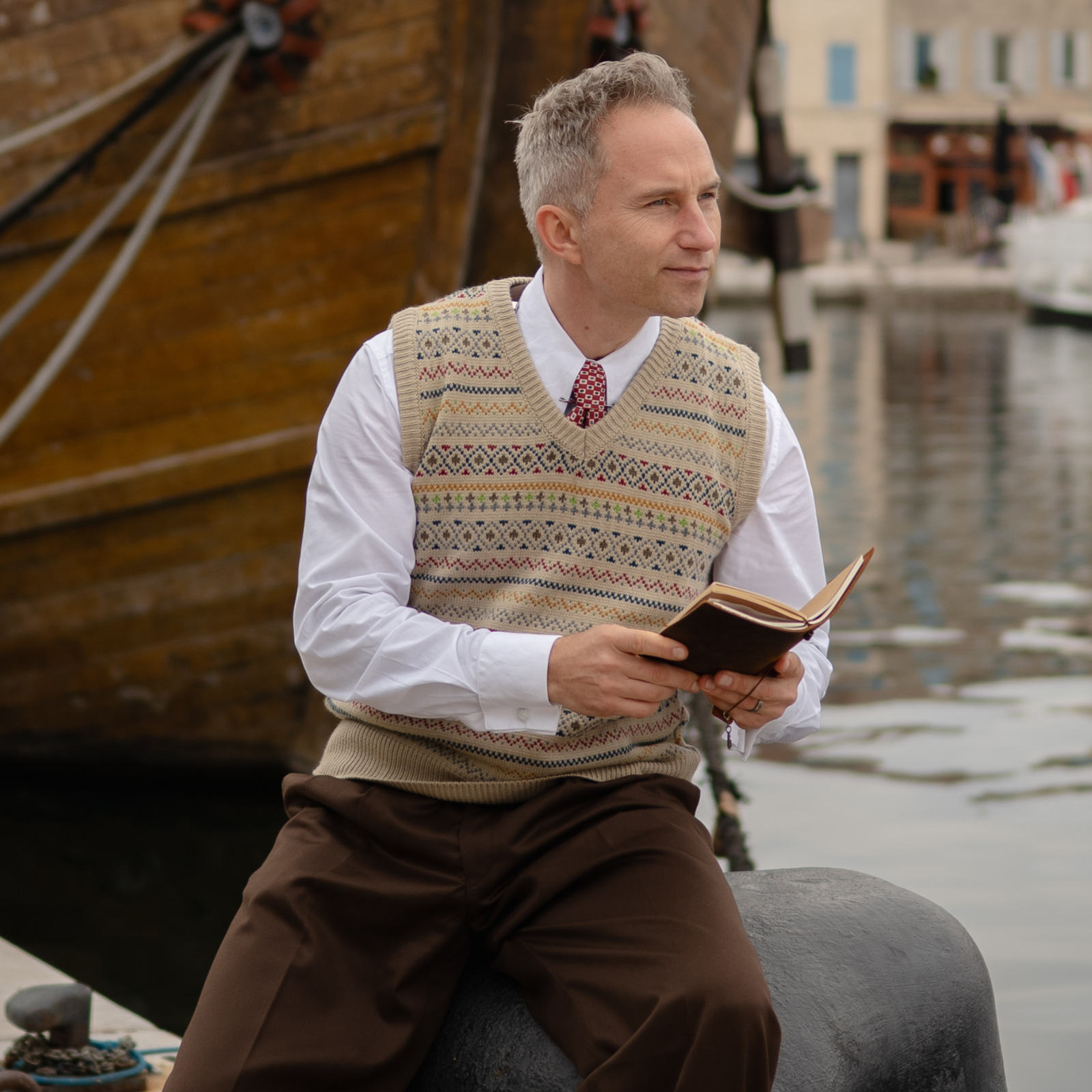 Man reading a book by a waterfront with a boat in the background