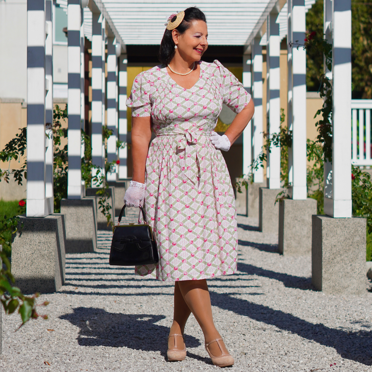 Model wears a 1940s-style pink and cream floral short-sleeve blouse with a sweetheart neckline, smocking detailing on shoulders and waist, and a waist tie.  
