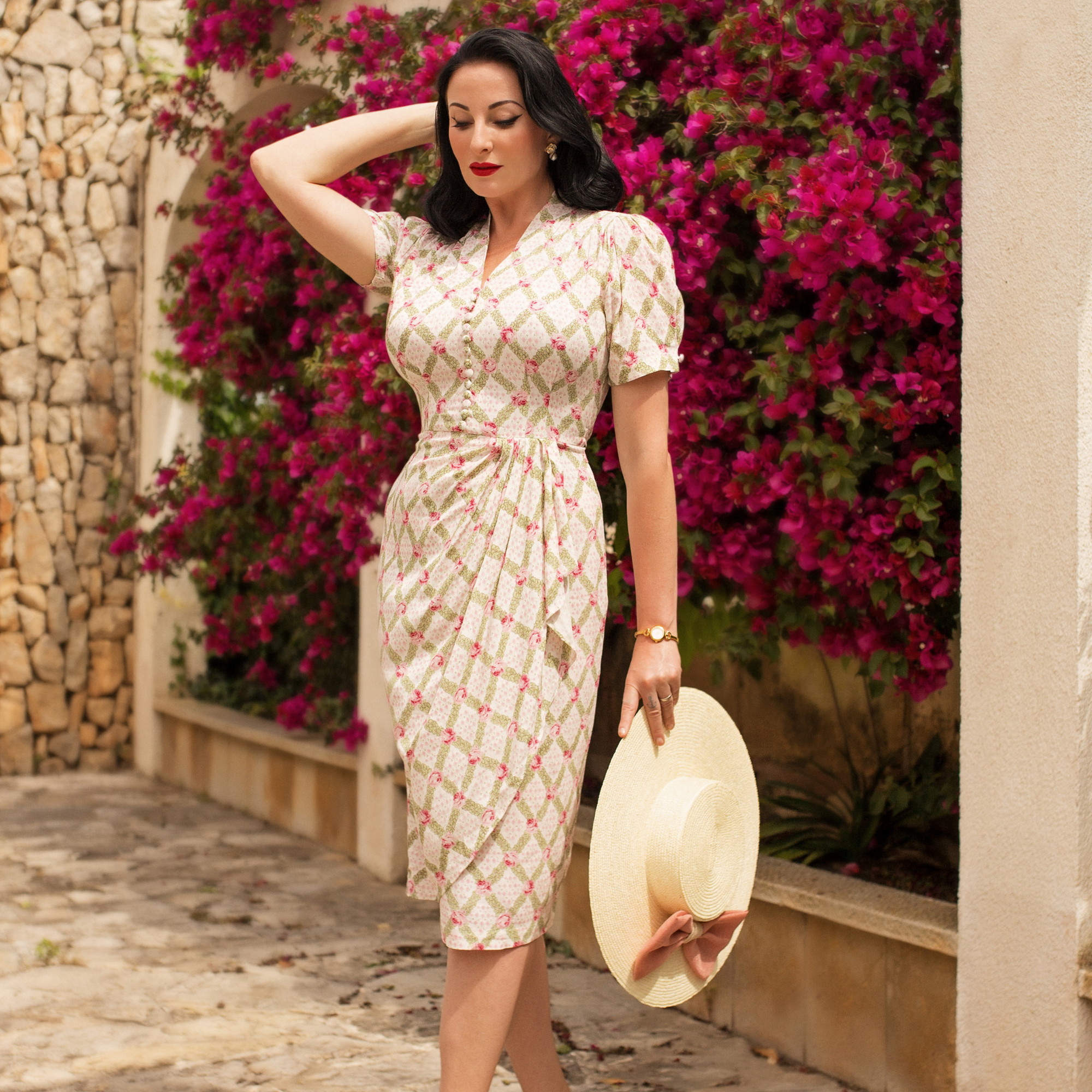 A woman wearing a rose kiss print dress with a fitted style, featuring a waterfall ruffle on the waist and decorative buttons on the front bodice. She is holding a hat and standing near a wall with pink flowers.