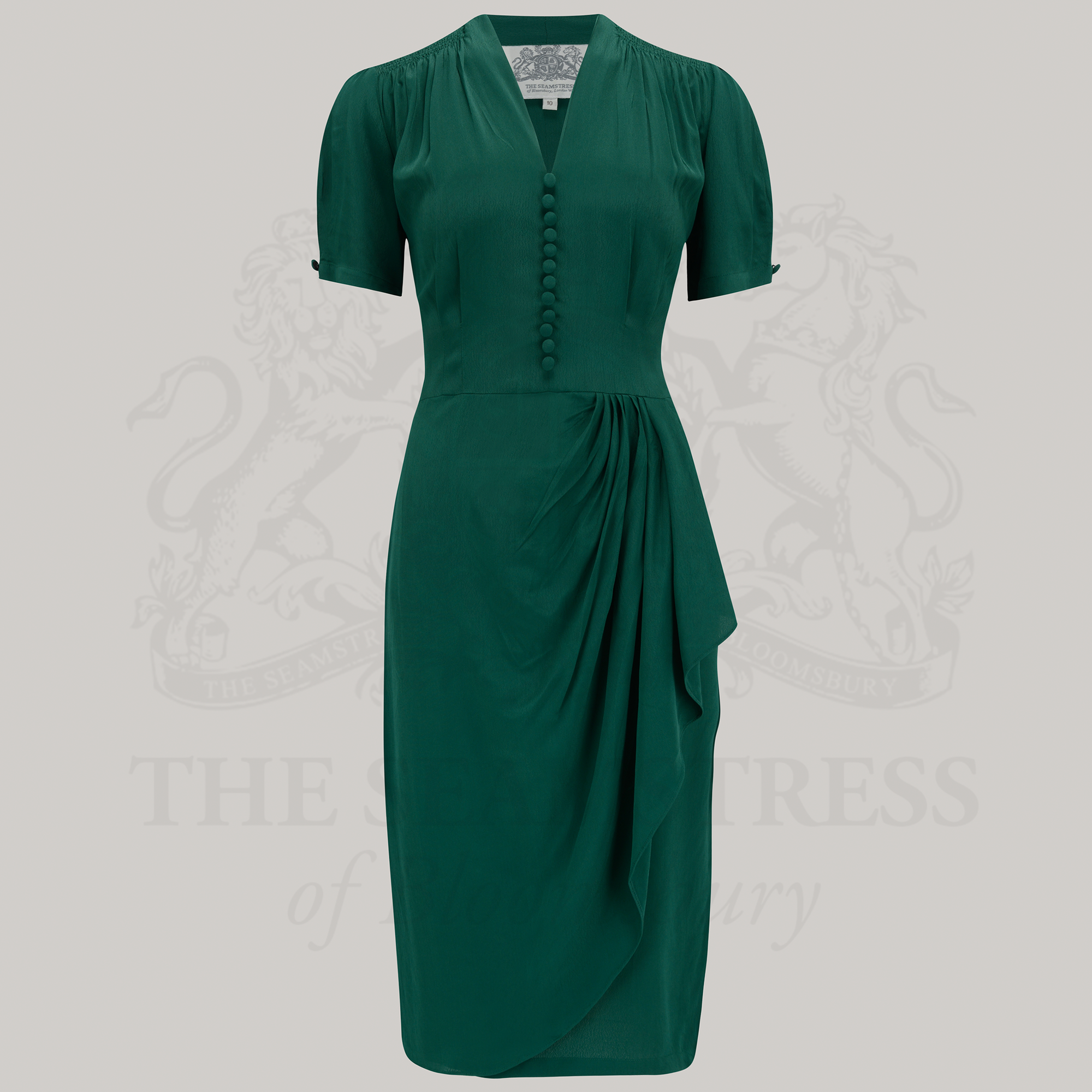 A green, short-sleeved Mabel Waterfall Dress with a waterfall ruffle on the waist and decorative buttons on the front bodice, pictured against a grey background.