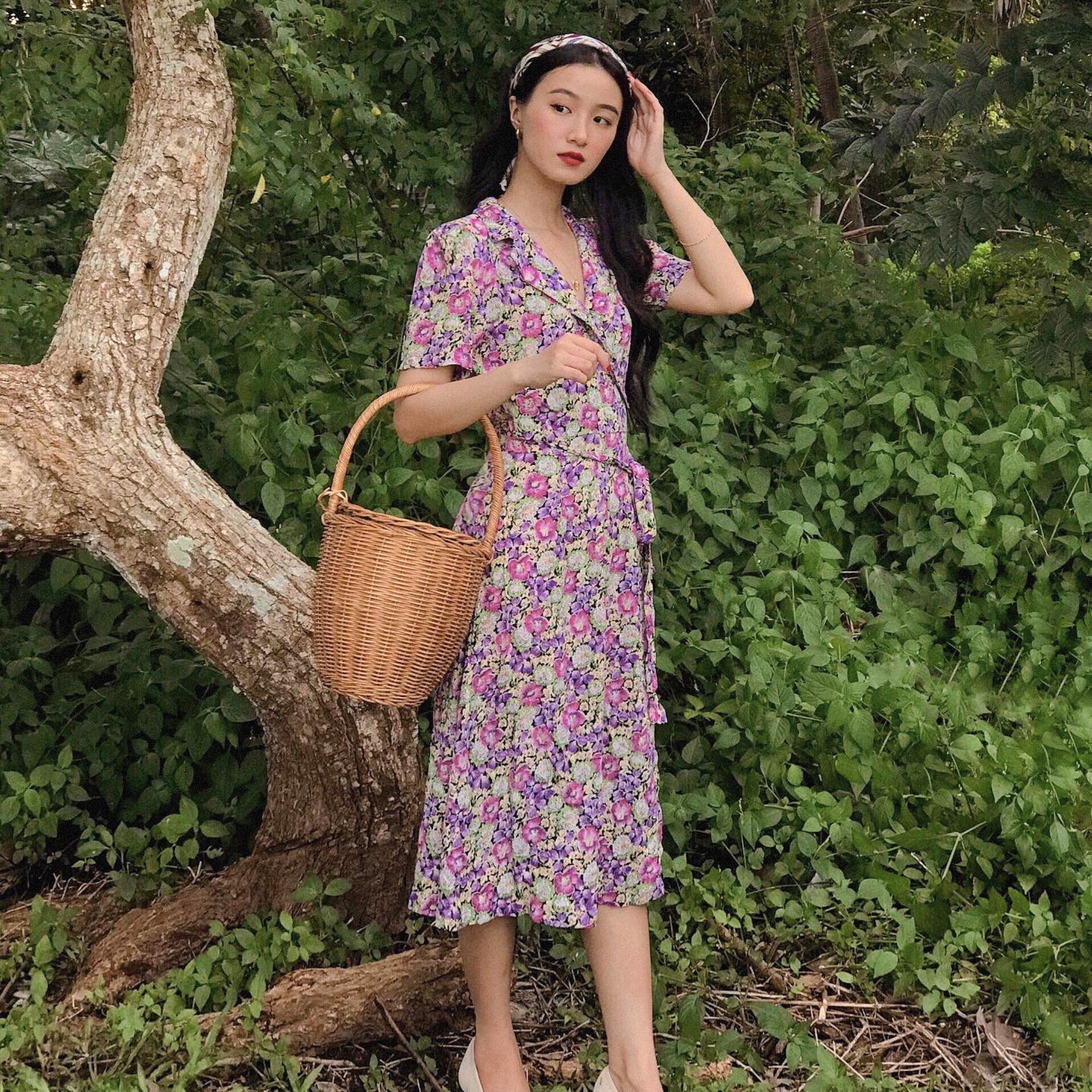 A woman stands outdoors in a lush green setting next to a large tree trunk, wearing a short-sleeved floral midi dress with a purple, pink, and green pattern. She is holding a brown wicker basket.