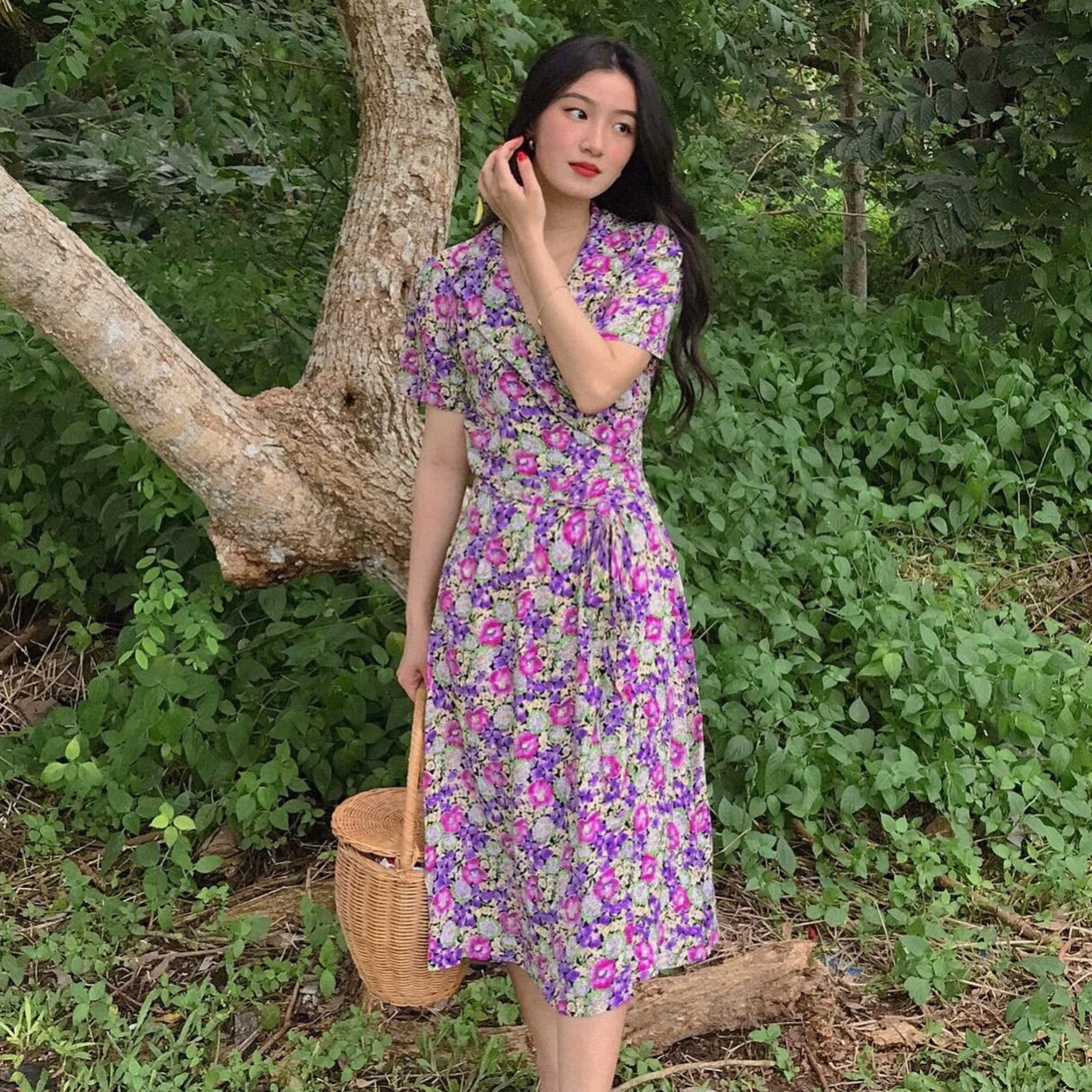A woman stands outdoors in a wooded area wearing a short-sleeved wrap-style midi dress with a dense floral print in purple, pink, and green. She is holding a woven basket.