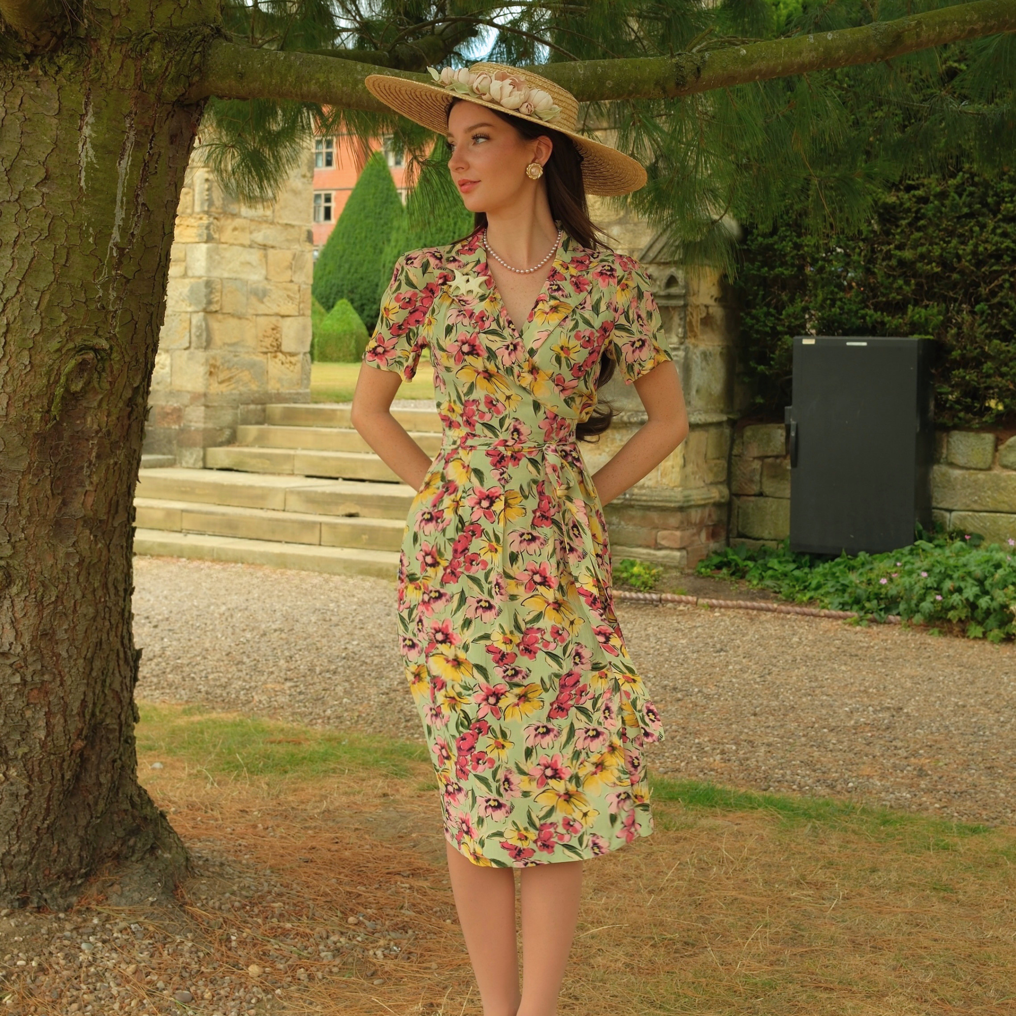 A woman wearing a short-sleeve knee-length floral dress and a wide-brimmed straw hat stands outdoors in a garden setting with a stone building and tree trunk in the background.