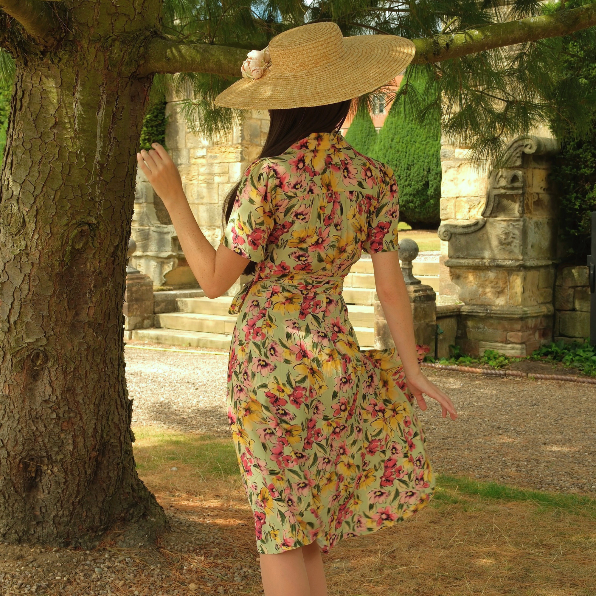 A woman wearing a short-sleeve floral wrap dress and a wide-brimmed straw hat stands outdoors next to a large tree, viewed from behind.