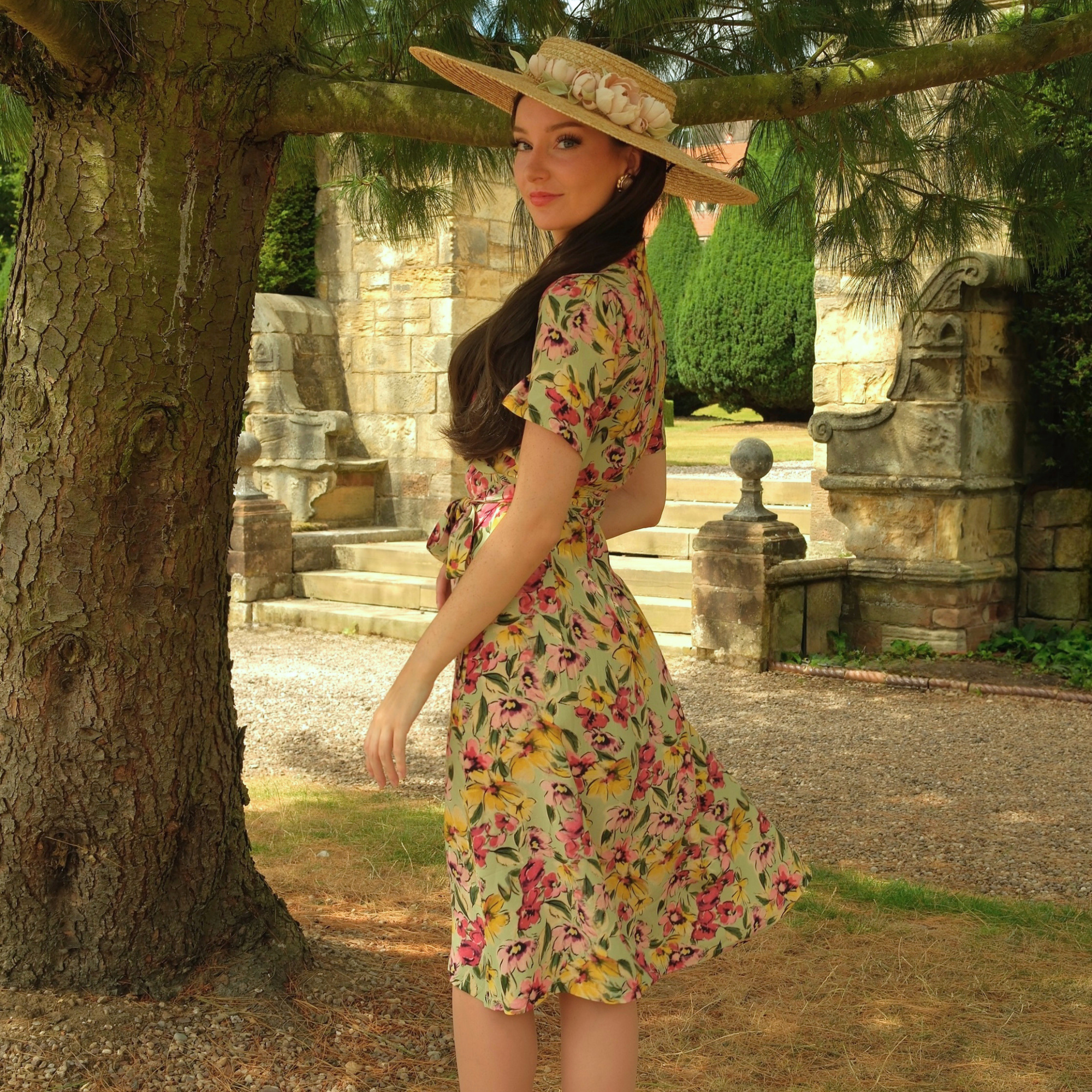 A woman wearing a short-sleeve floral dress and a wide-brimmed straw hat stands outdoors in front of a stone garden structure and large tree trunk.