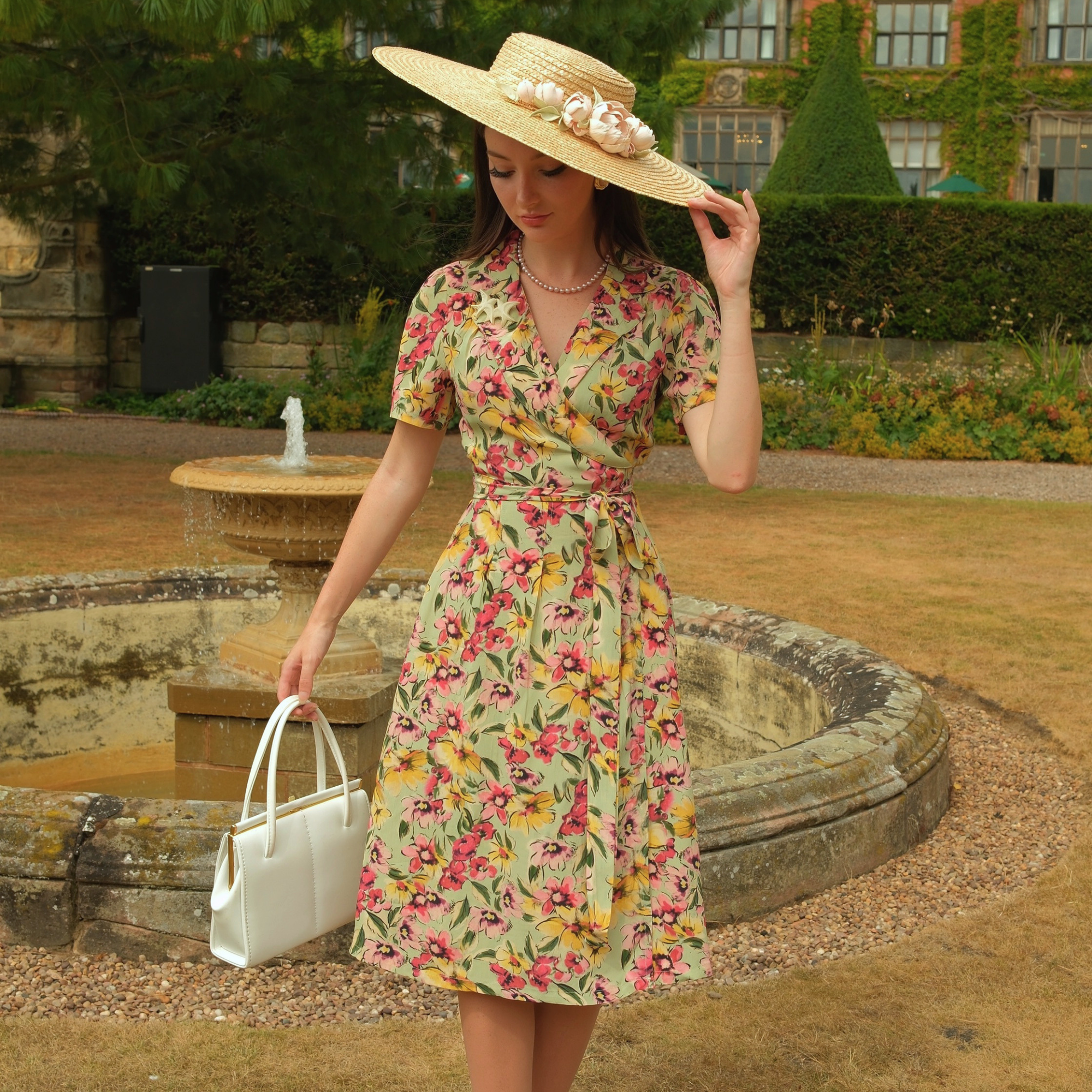A woman stands in a garden courtyard wearing a short-sleeve wrap-style midi dress with a colorful floral print, a wide-brim straw hat with flowers, and holding a white handbag.