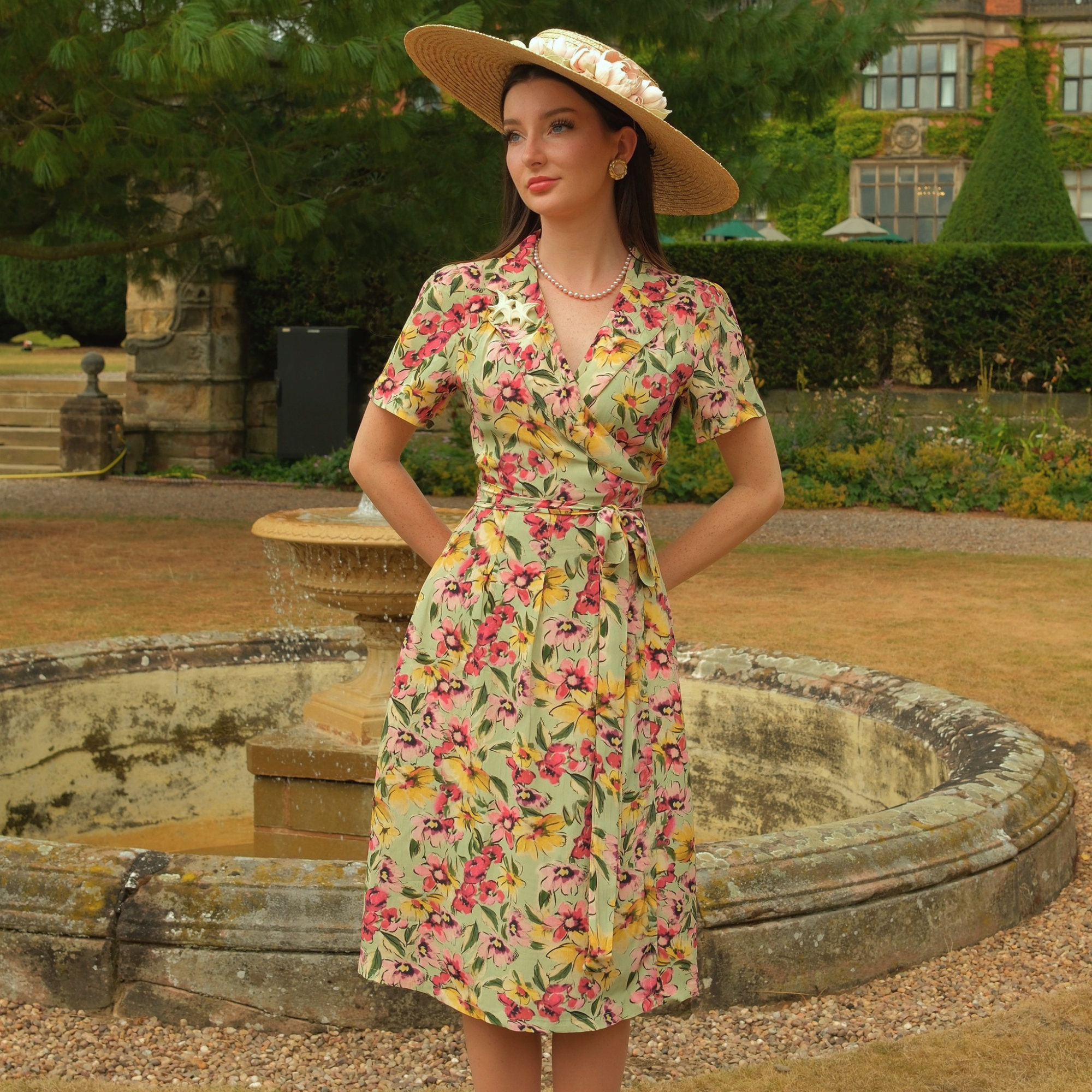 A woman stands outdoors in front of a stone fountain and manicured garden hedges, wearing a short-sleeve wrap dress with a floral print and a wide-brim straw hat.