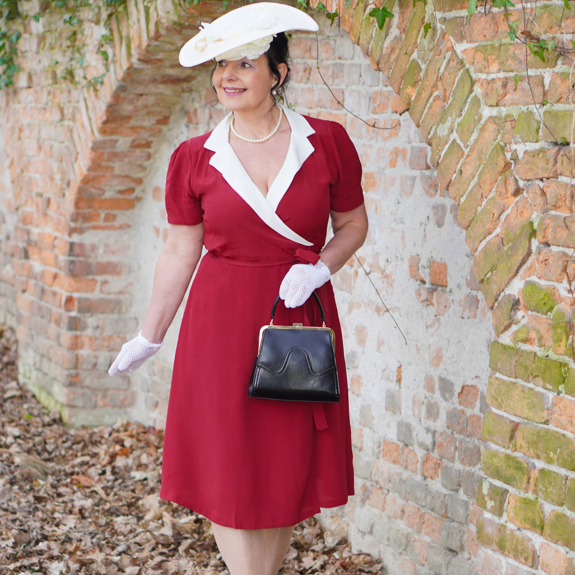 A woman wearing a red and ivory vintage-style wrap dress with a tie at the waist, V-neck, and short sleeves. She is accessorised with white gloves, a white hat, and a black purse.