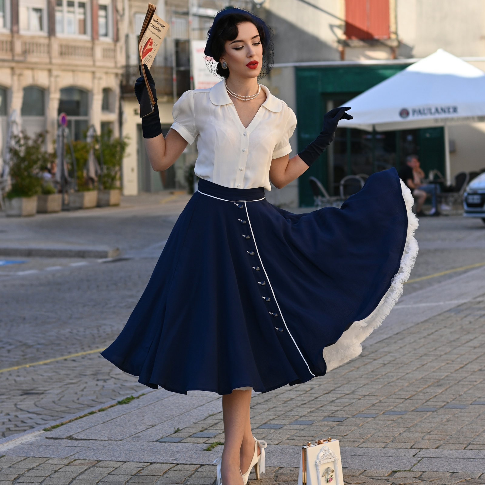 A woman wearing a full navy swing skirt with cream contrasting elements and buttons, paired with a white top, gloves, and heels, standing on a cobblestone street.