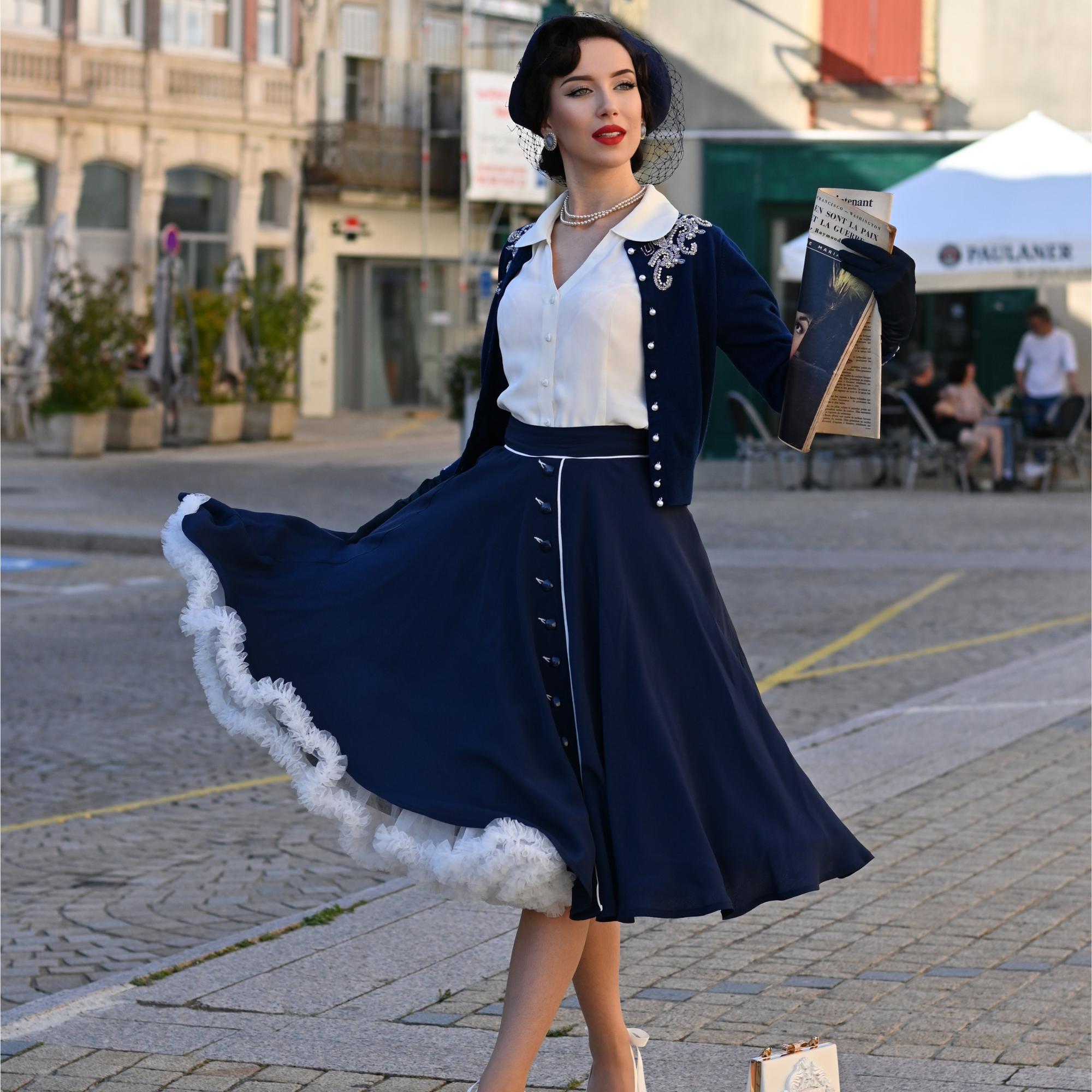 A woman wearing a full navy swing skirt with cream contrasting elements and buttons, paired with a white top, gloves, and heels, standing on a cobblestone street.