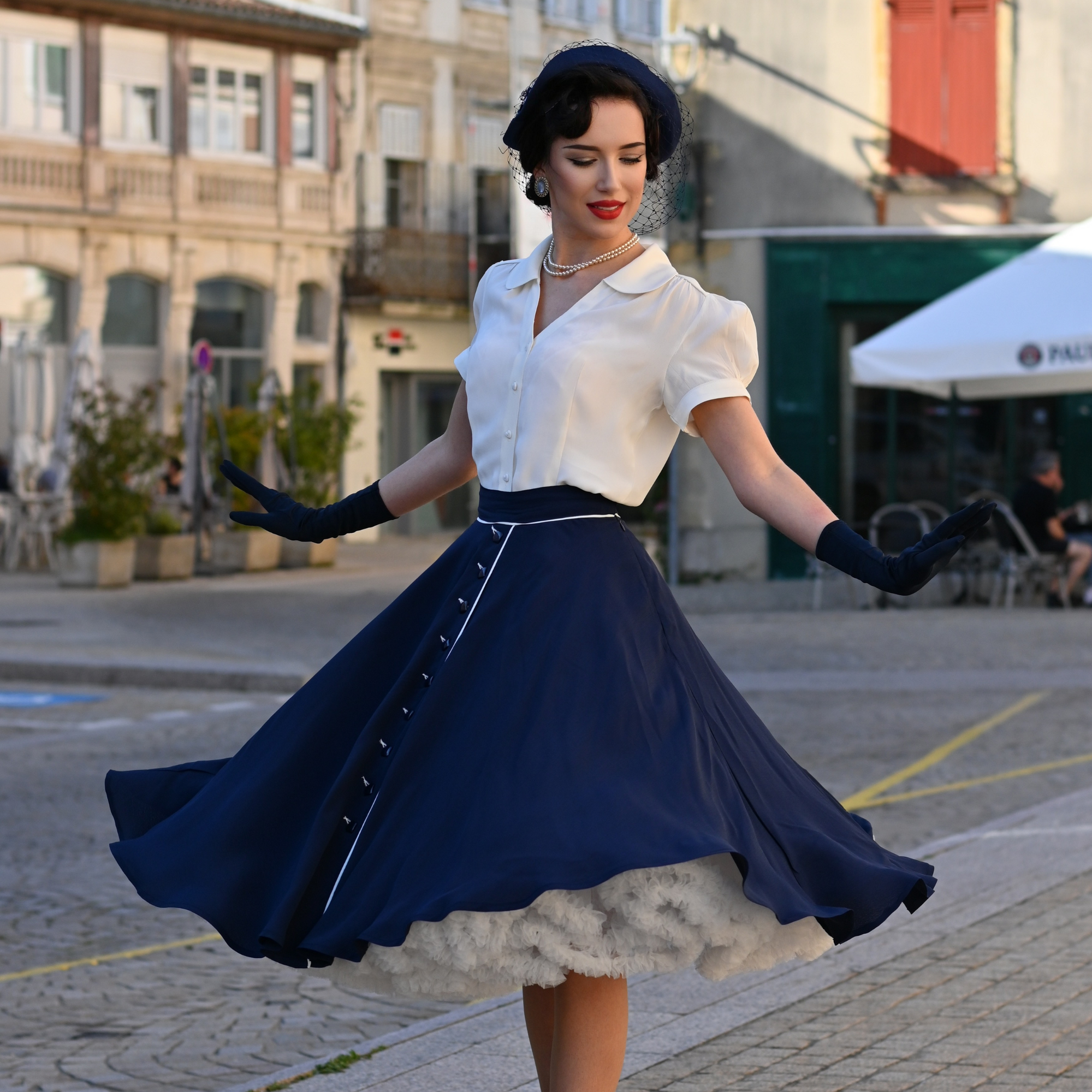 A woman wearing a full navy swing skirt with cream contrasting elements and buttons, paired with a white top, gloves, and heels, standing on a cobblestone street.