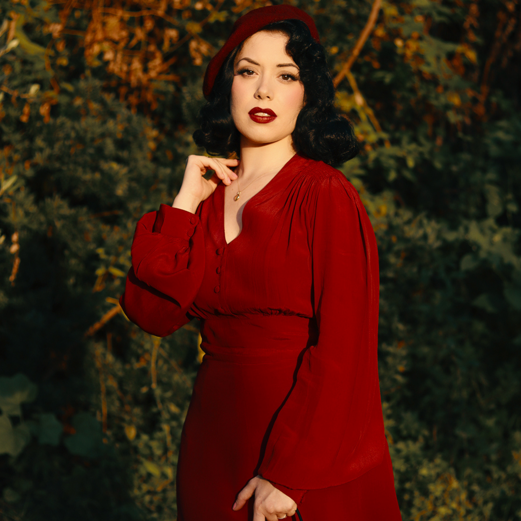 Model wears a 1940s style long sleeve dress in dark red/burgundy. Featuring gauged-in top yoke detailing on the shoulders. 4 small buttons on the bodice, and a tie-waist belt.