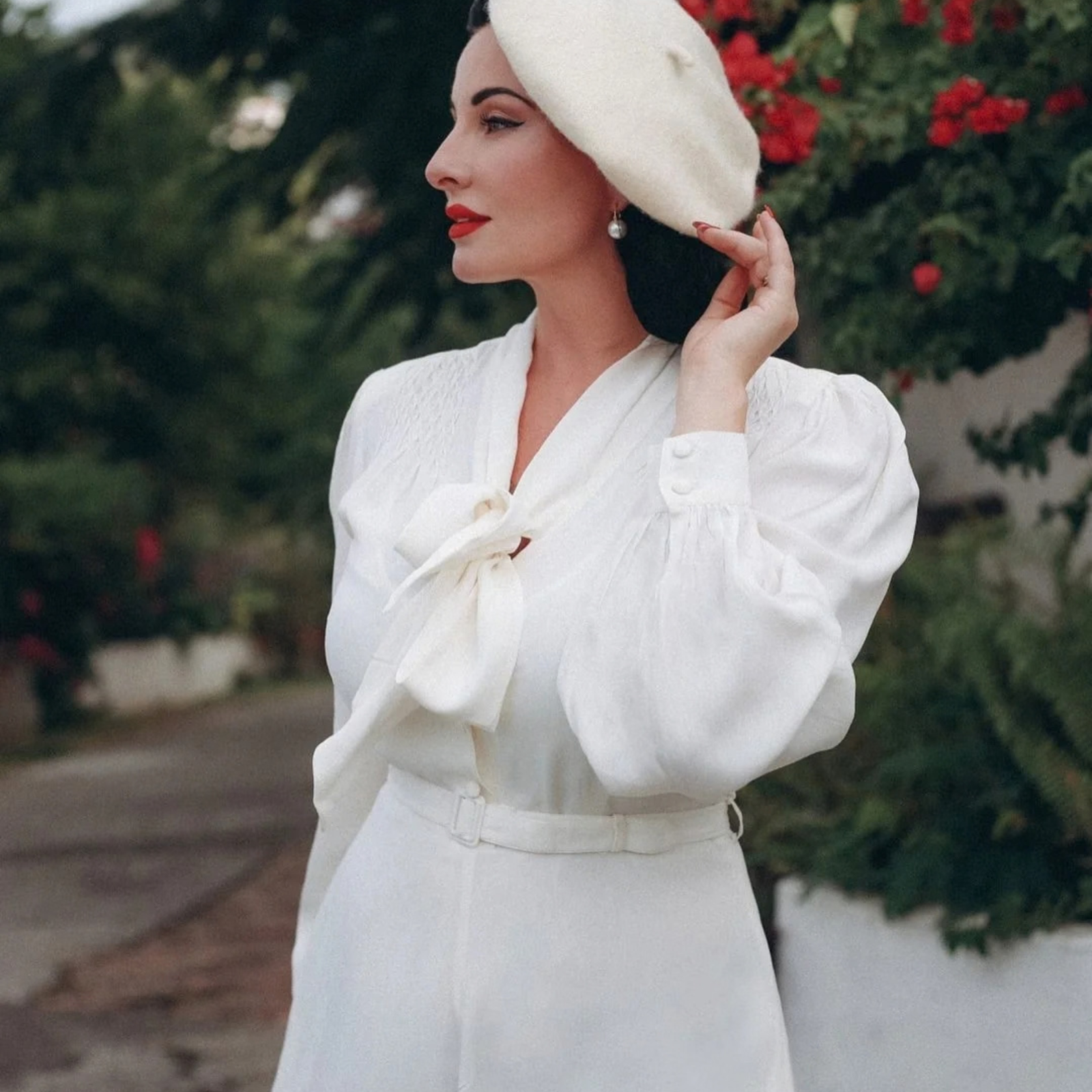 A woman wearing a white long-sleeve dress with a large bow at the neckline, a matching belt, and a white beret, standing outdoors in front of green foliage and red flowers.