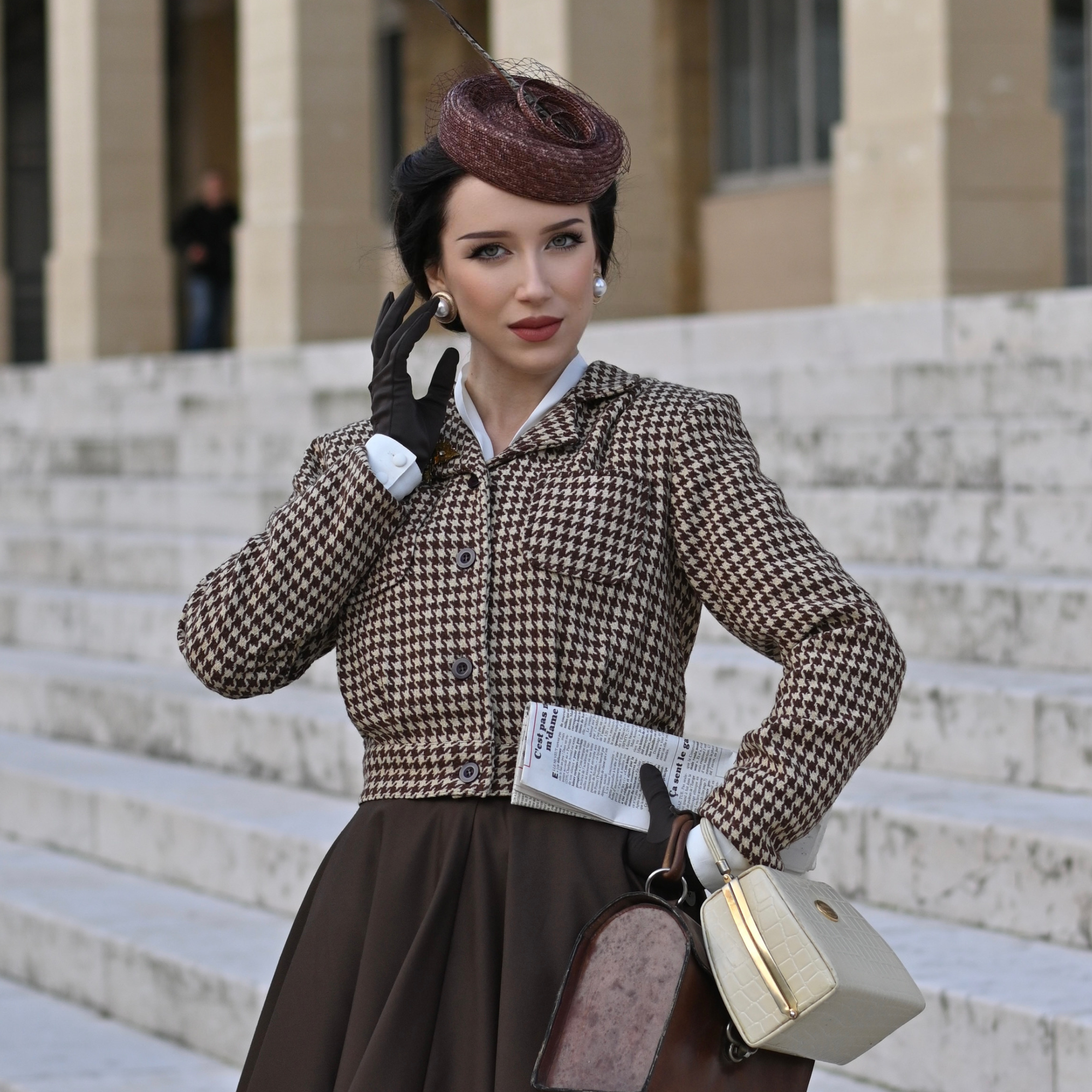 Woman in vintage-style outfit with houndstooth jacket and brown hat, holding a suitcase and newspaper, standing on steps.