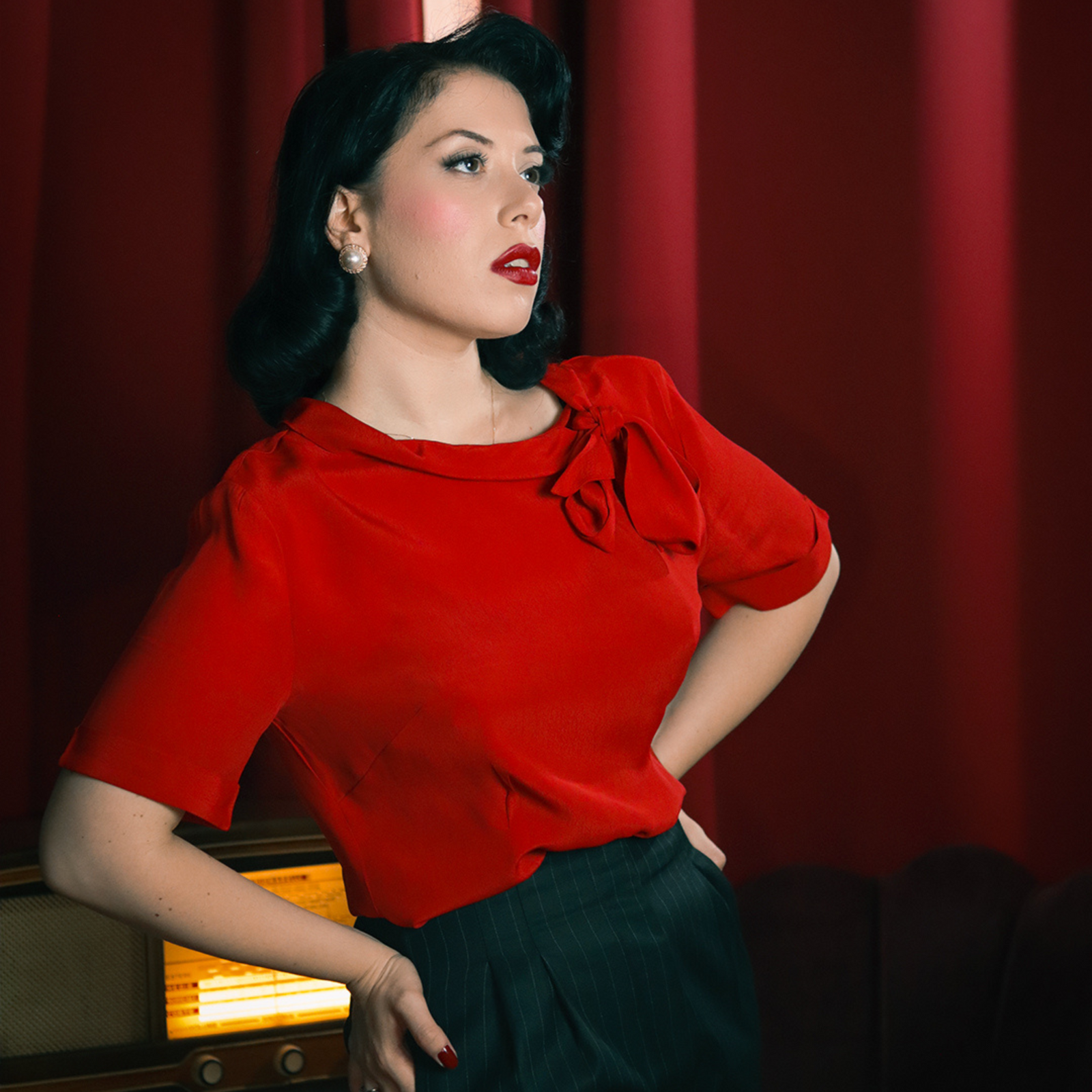 Model wears a red short sleeve 1940s-style blouse with side bow collar, paired with black pinstriped trousers. 