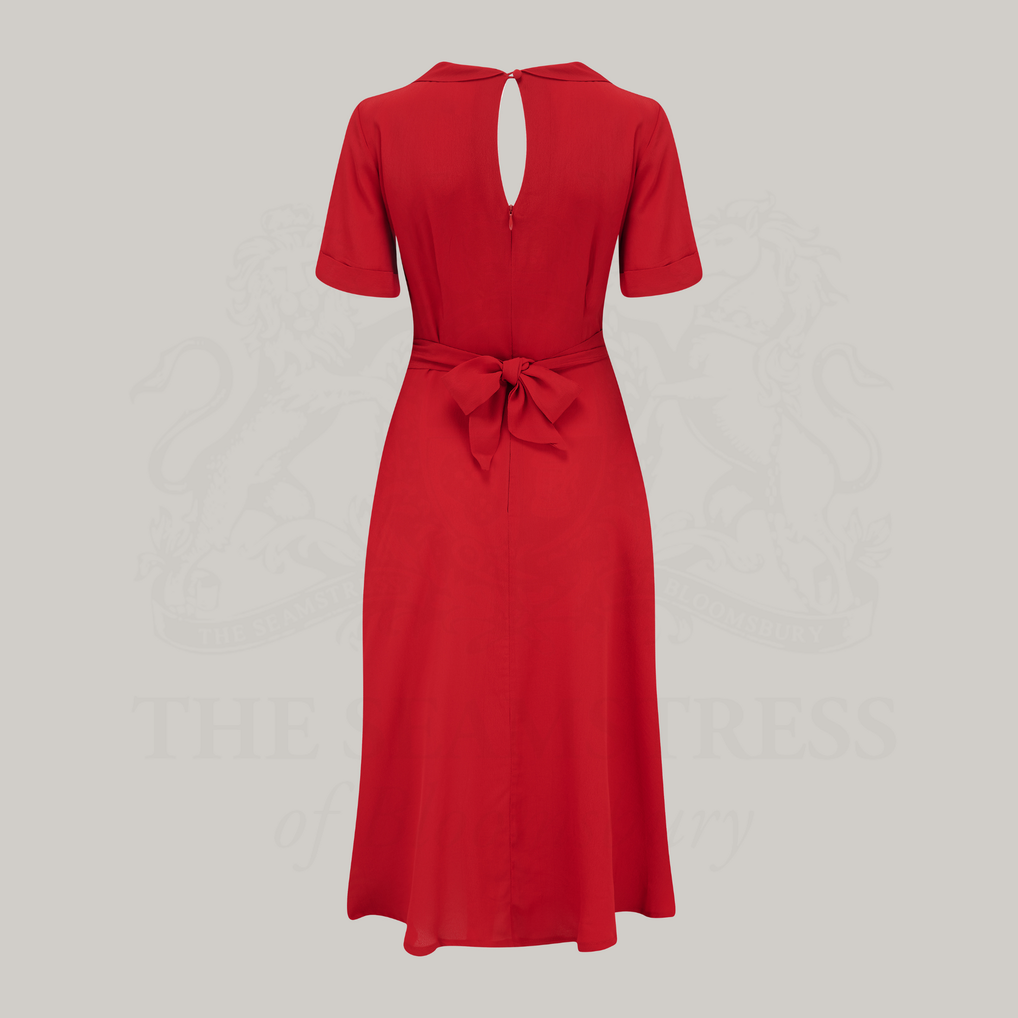 A 1940s vintage style short-sleeved dress with a side tie-neck and a tie-waist belt. This dress is red, has an a-line skirt, and is designed to finish below the knee.
