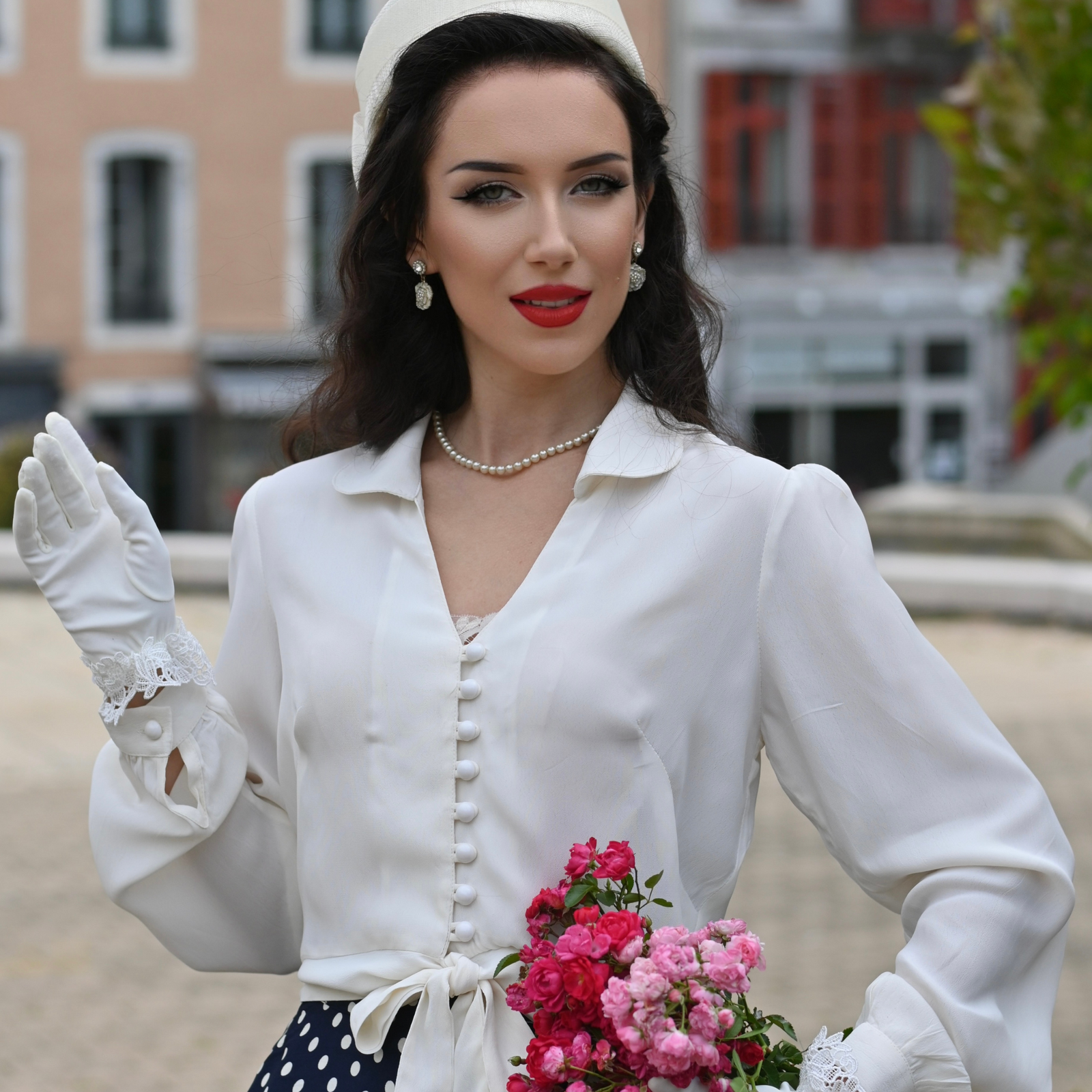 Model wears a 1940s-style cream circle long-sleeve blouse with a v-neck, button fastenings on the front, and a waist tie.
