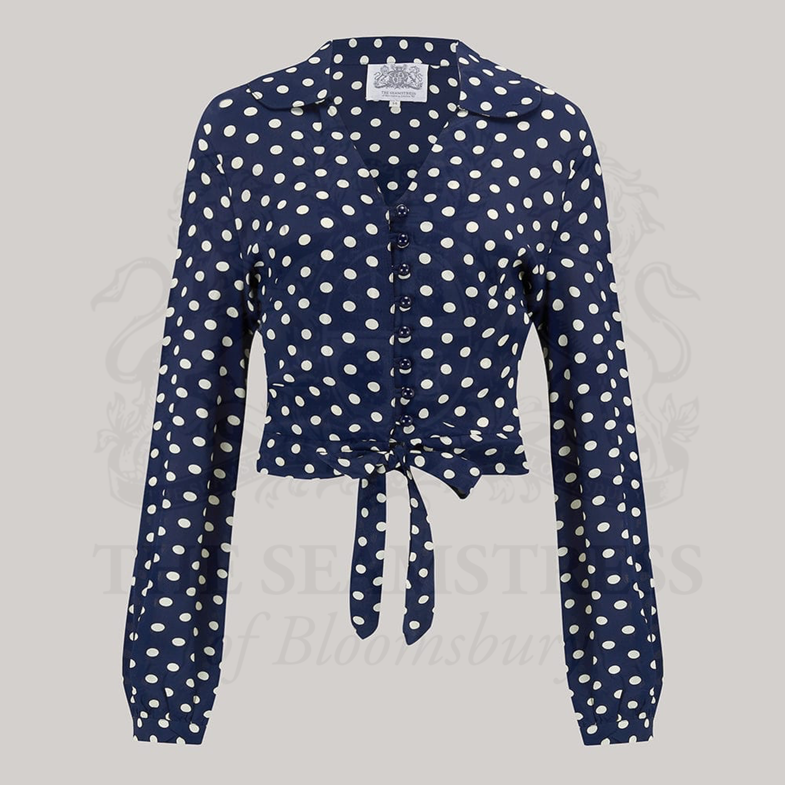 A 1940s-style long-sleeve blouse in navy with a cream polka dot pattern. The blouse has a v-neck, button fastenings on the front, and a waist tie.
