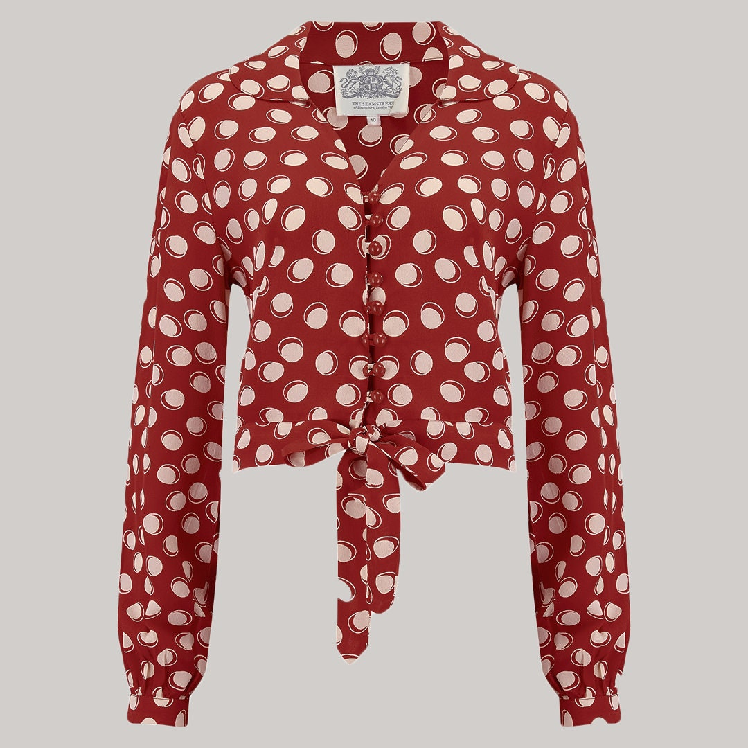 A red long-sleeve blouse with a cream polka-dot pattern, featuring a button-up front, a collar, and a tie-front waist. 