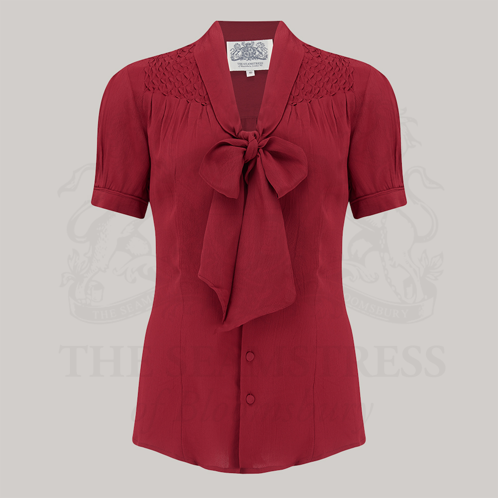A short-sleeved red blouse with a pussy-bow neckline and smock stitching on the shoulders, button fastenings, and a collared neckline.