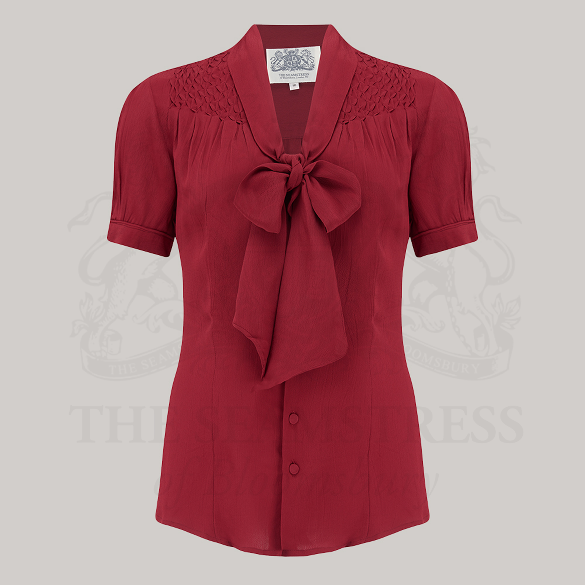 A short-sleeved red blouse with a pussy-bow neckline and smock stitching on the shoulders, button fastenings, and a collared neckline.