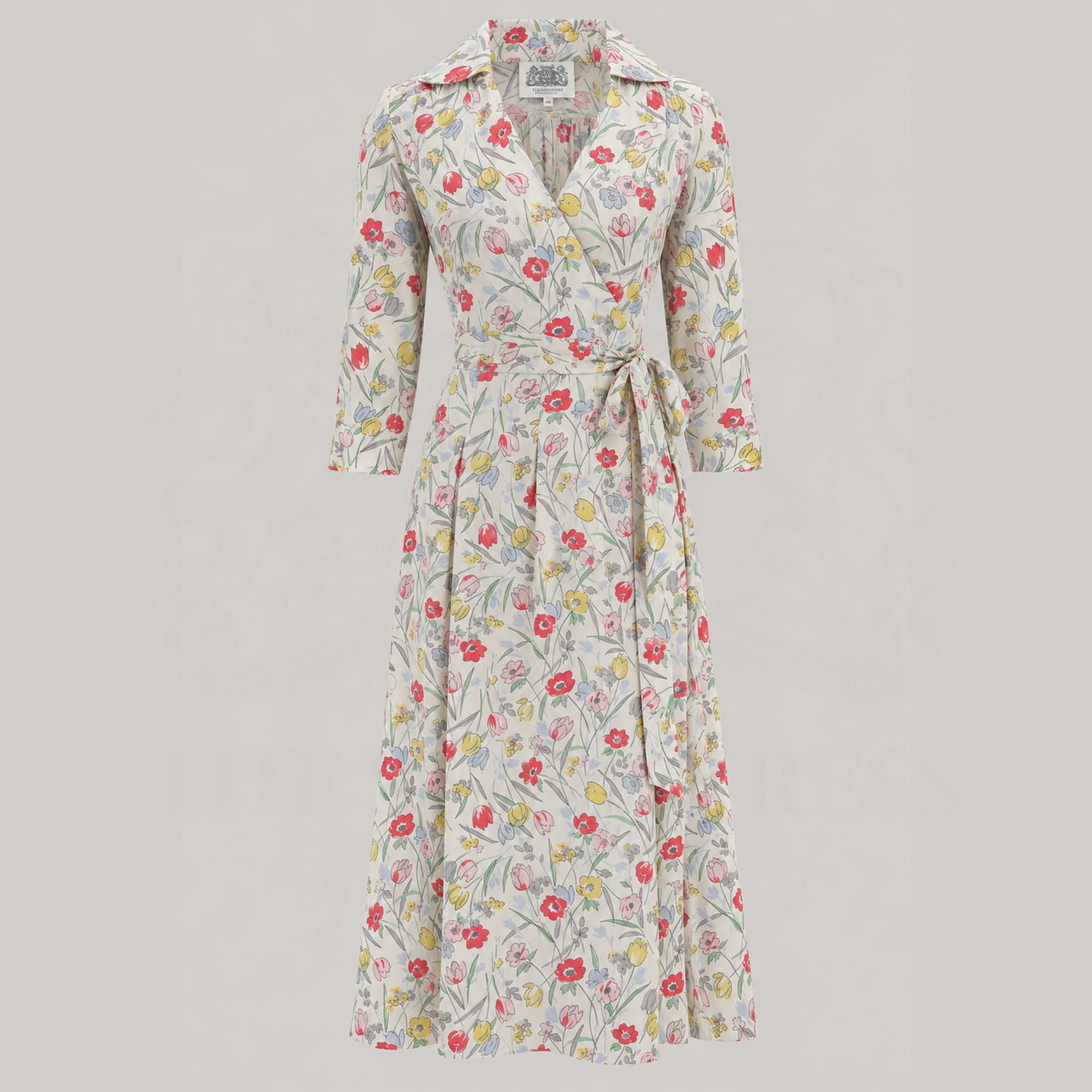 A long-sleeve wrap dress with a tie waist, featuring an all-over floral print in red, yellow, and green on a light beige background, shown against a solid light grey studio backdrop.