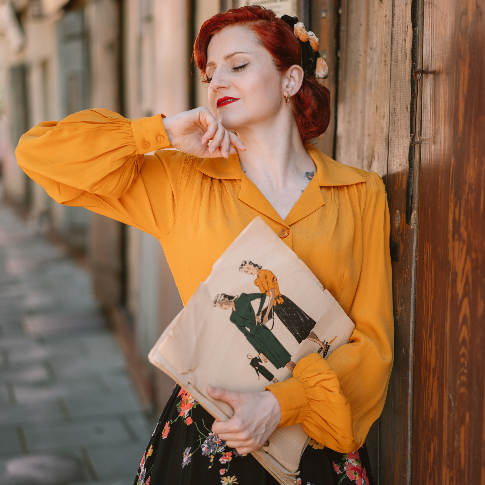 A woman wearing a mustard yellow blouse with a revere collar and long bishop sleeves, paired with a floral skirt, standing in front of a wooden door.