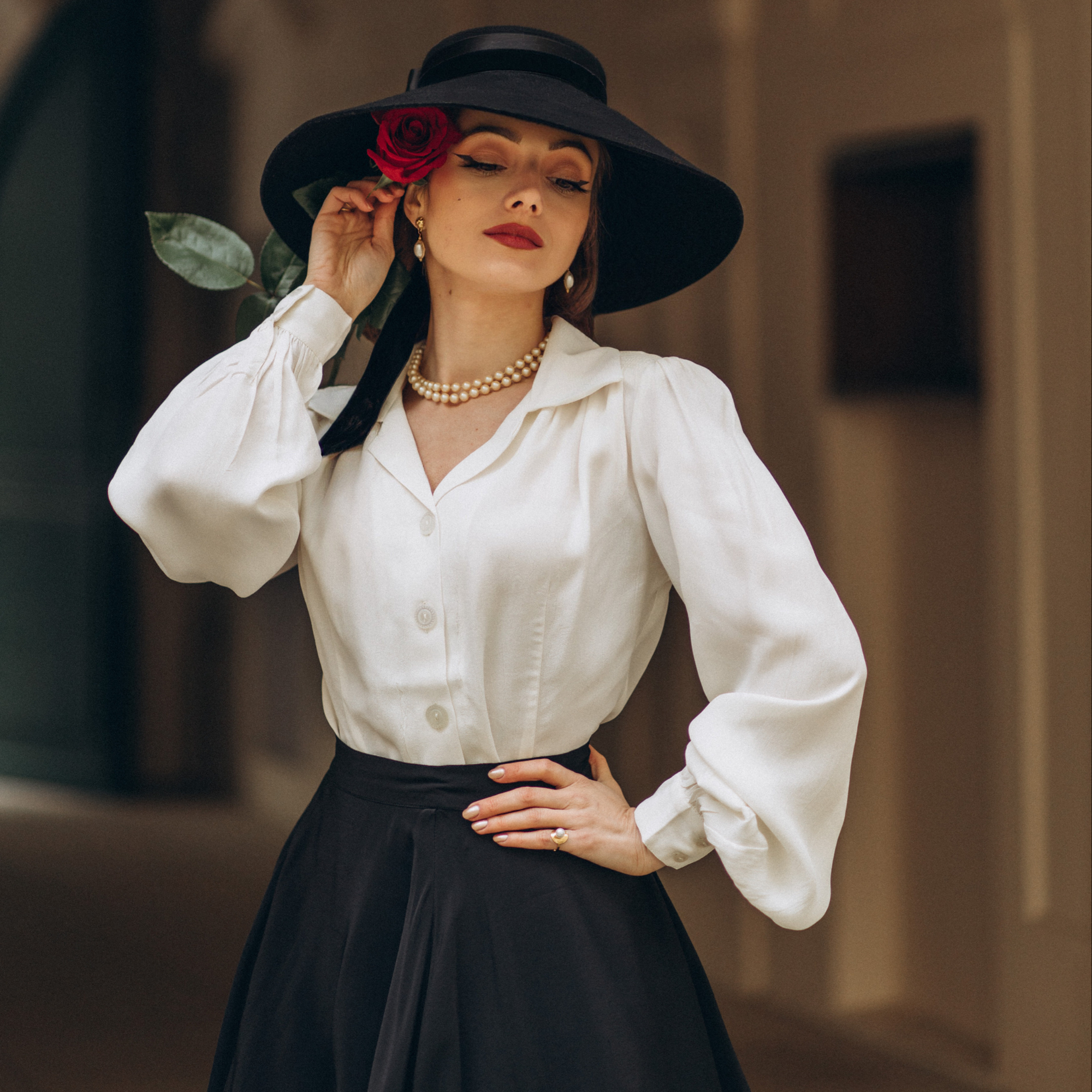 A woman wearing a vintage style cream blouse with a revere collar, long bishop sleeves with full cuffs, and shoulder pads. She is also wearing a black skirt, black hat with a red rose, and pearls.