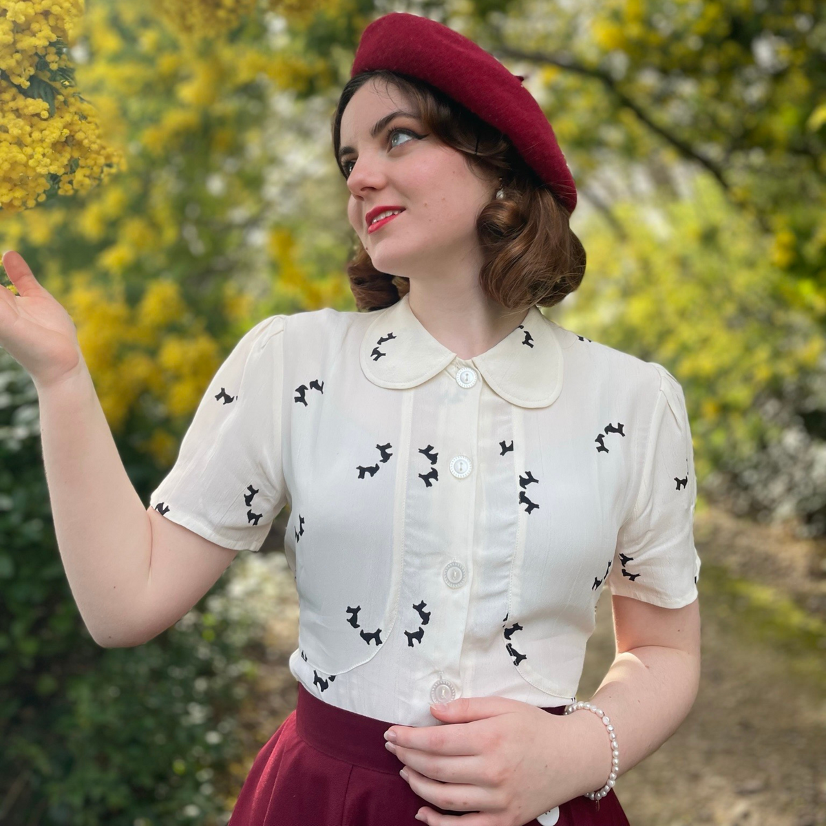 Model wears a 1940s style short sleeve blouse and a burgundy beret. The blouse is cream coloured with small black dogs dotted around the blouse. Featuring a Peter-Pan front collar, buttons down the front, and a tie-waist belt. 