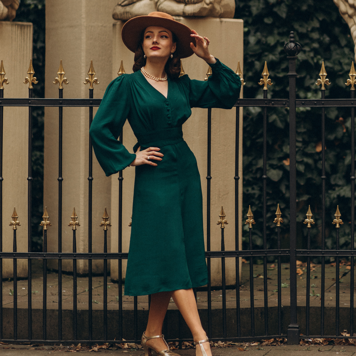 Model wears a 1940s style long sleeve dress in dark green with a brown wide brim hat and pearl jewellery. Featuring gauged-in top yoke detailing on the shoulders and sleeves that puff out fully at the cuff. 4 small buttons down the front of the dress, and a tie-waist belt. 