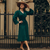 Model wears a 1940s style long sleeve dress in dark green with a brown wide brim hat and pearl jewellery. Featuring gauged-in top yoke detailing on the shoulders and sleeves that puff out fully at the cuff. 4 small buttons down the front of the dress, and a tie-waist belt. 