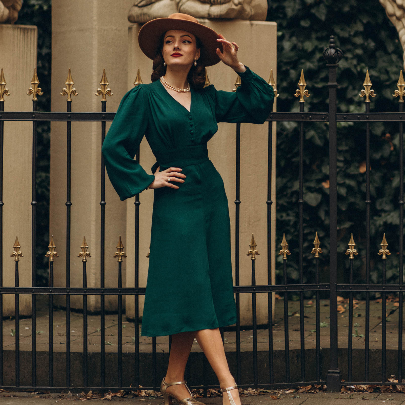 Model wears a 1940s style long sleeve dress in dark green with a brown wide brim hat. Featuring gauged-in top yoke detailing on the shoulders. 4 small buttons on the bodice, and a tie-waist belt. 