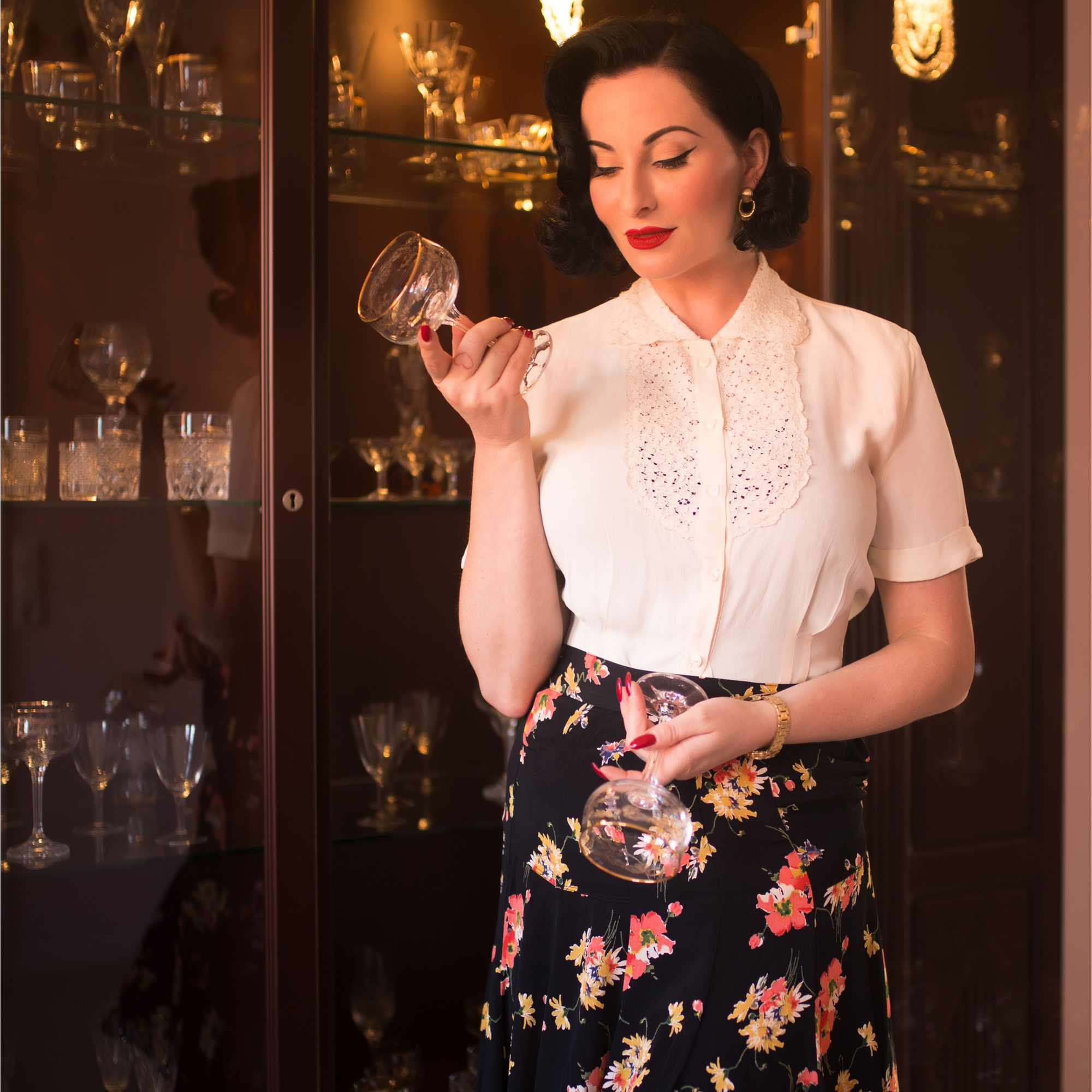 Model wears 1940s black dancing skirt with floral pattern paired with a cream blouse which has lace detailing around the collar and chest. 