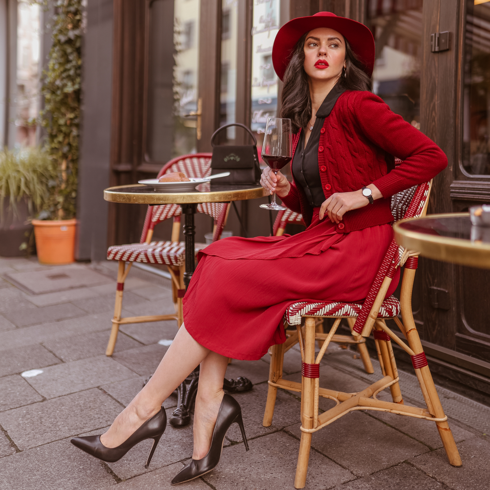 A model sits outside a bistro wearing a 1940s-style outfit, featuring a midi-skirt, blouse, and cardigan.