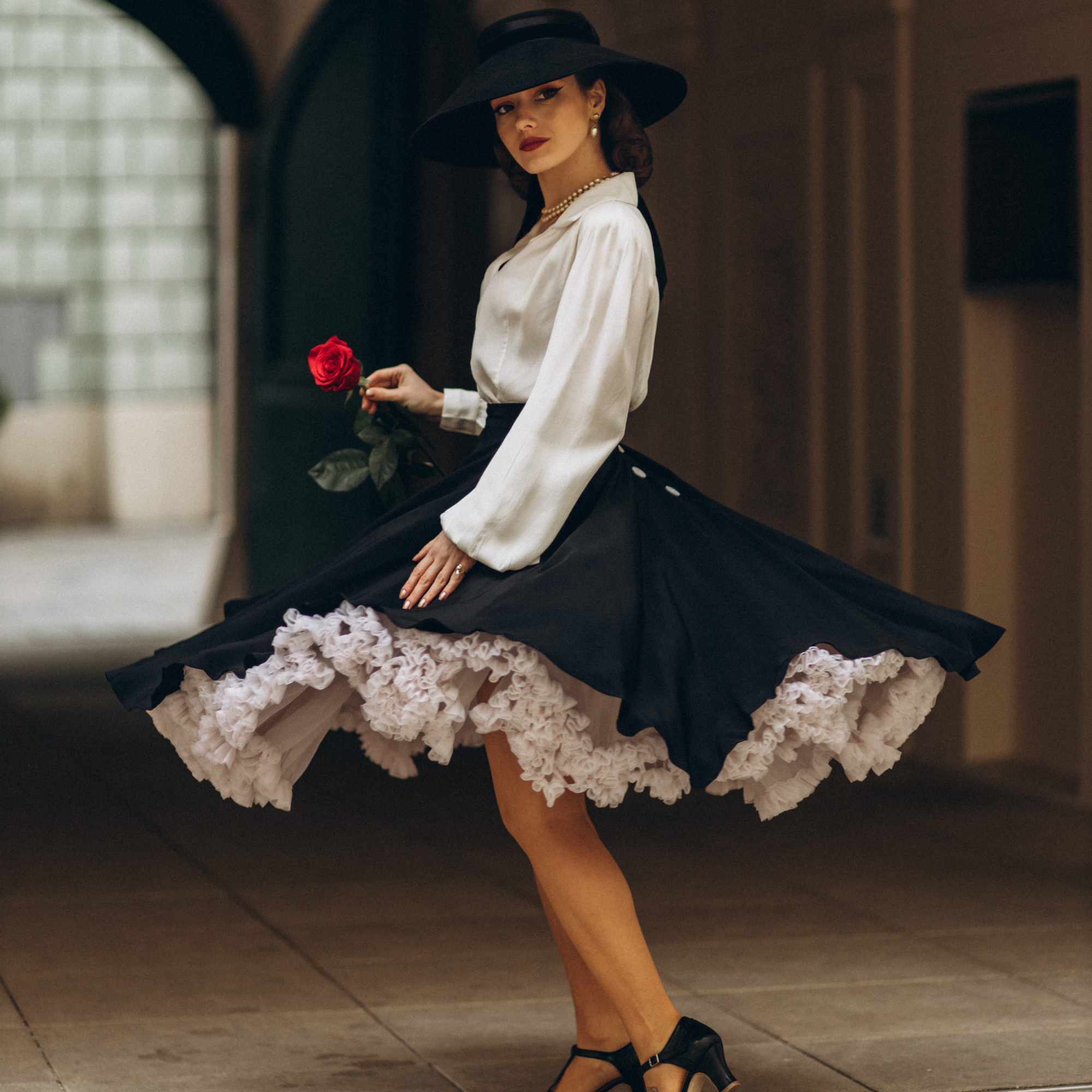 A woman wearing a black swing dance skirt with a full-circle silhouette, paired with a white blouse and a black hat. She is holding a red rose and dancing.