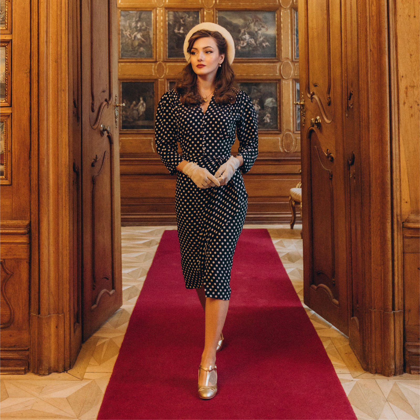 A woman standing in a room with wooden paneling and a red carpet, wearing a navy polka dot dress with 3/4 length sleeves, side zip closure, side seam pockets, top shoulder smocking, and a waterfall ruffle design.
