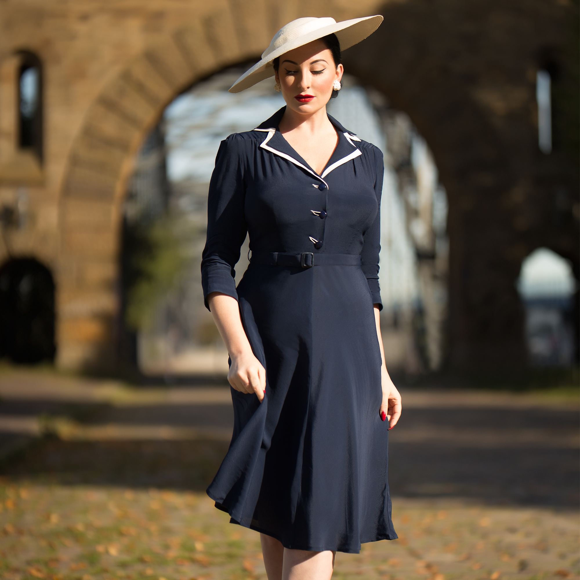A woman wearing a navy dress with a contrasting cream undercollar, square jet-style buttons, and a side zip closure. She is also wearing a wide-brimmed hat.