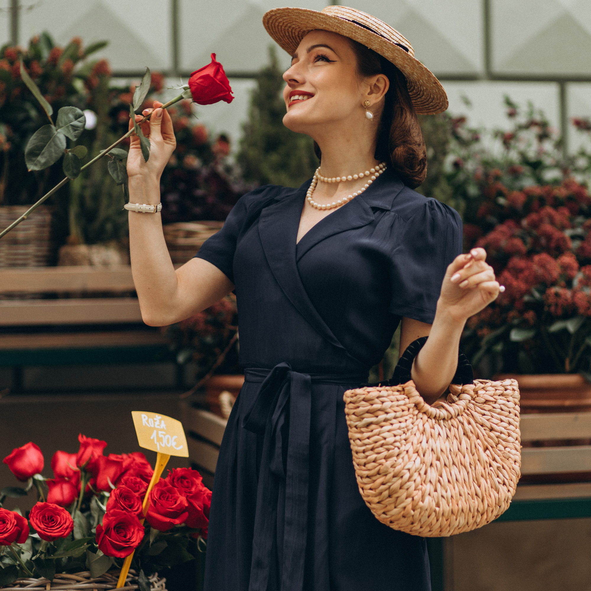A woman in a navy blue wrap dress with short sleeves and a v-neck, wearing a wide-brimmed hat, standing in a floral setting with red roses. She has side-seam pockets and is holding a woven tote bag.